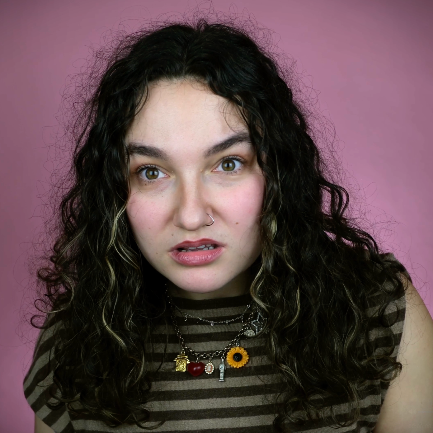 A young woman with curly dark hair, wearing a striped top and layered necklaces with charms, in front of a pink background.