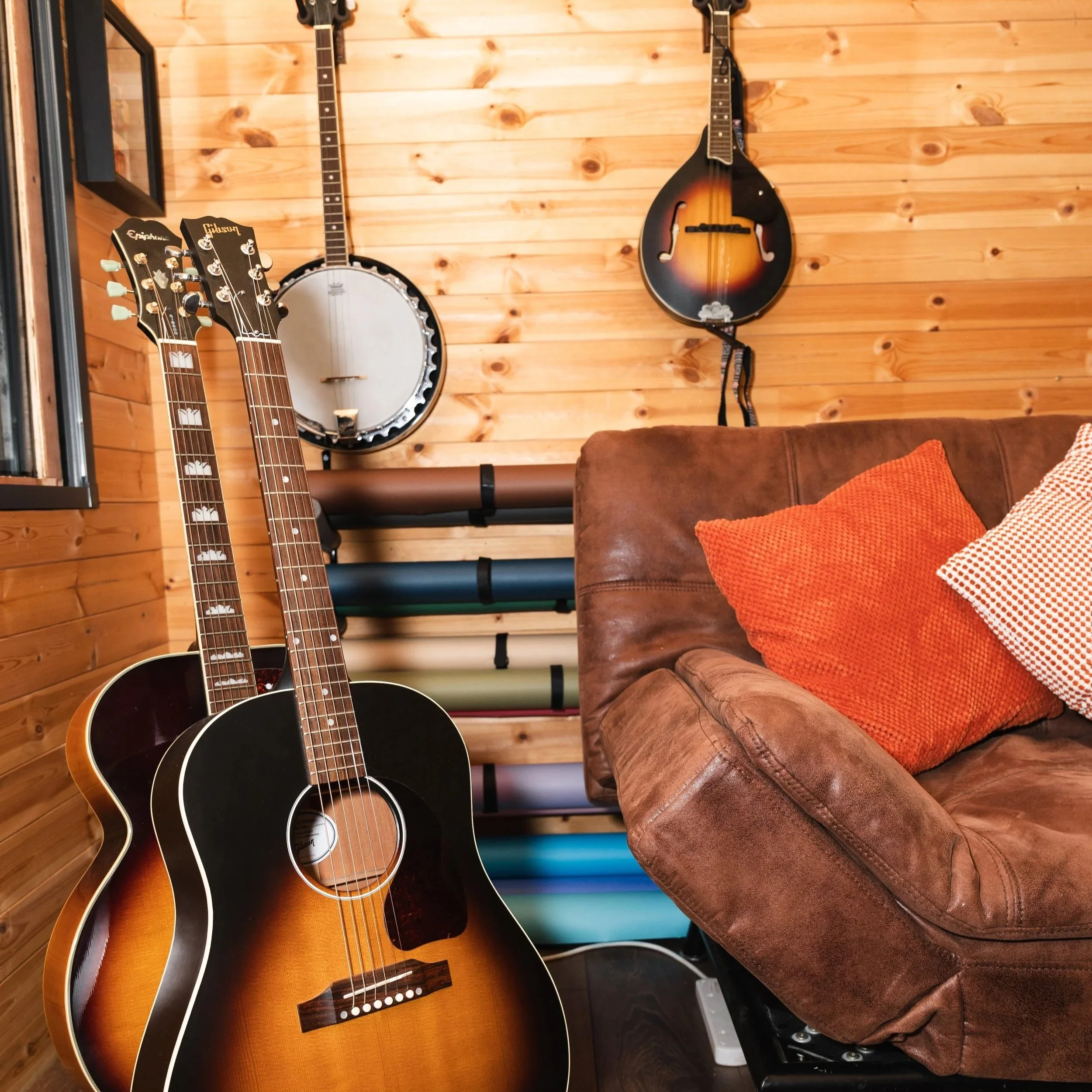 A cozy room with two acoustic guitars, a banjo, and a mandolin against a wooden wall, with a brown leather couch and textured pillows.