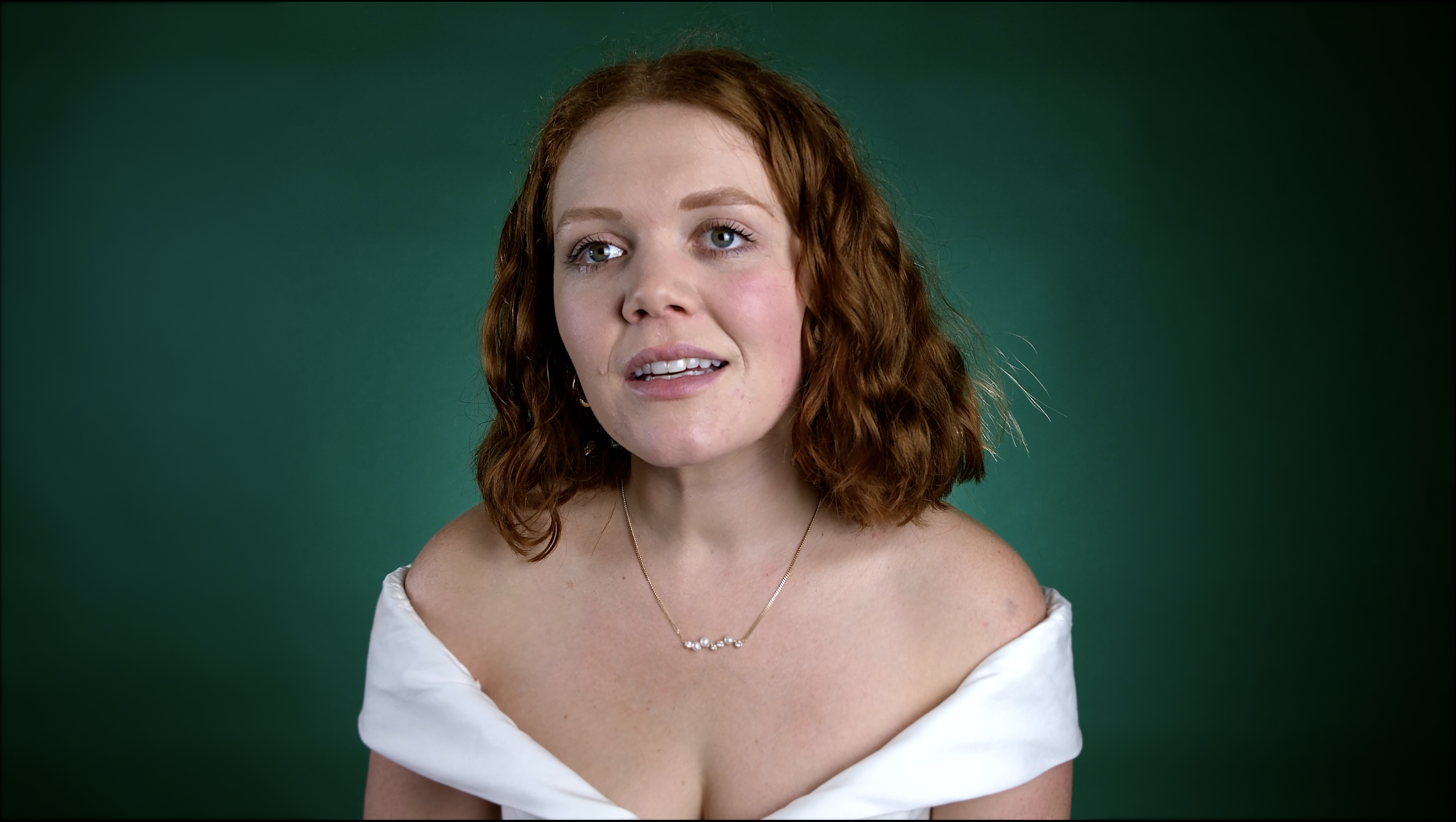 Close-up of a woman with red, wavy hair, wearing a white off-shoulder top and a delicate silver necklace, looking to her right against a solid green background.