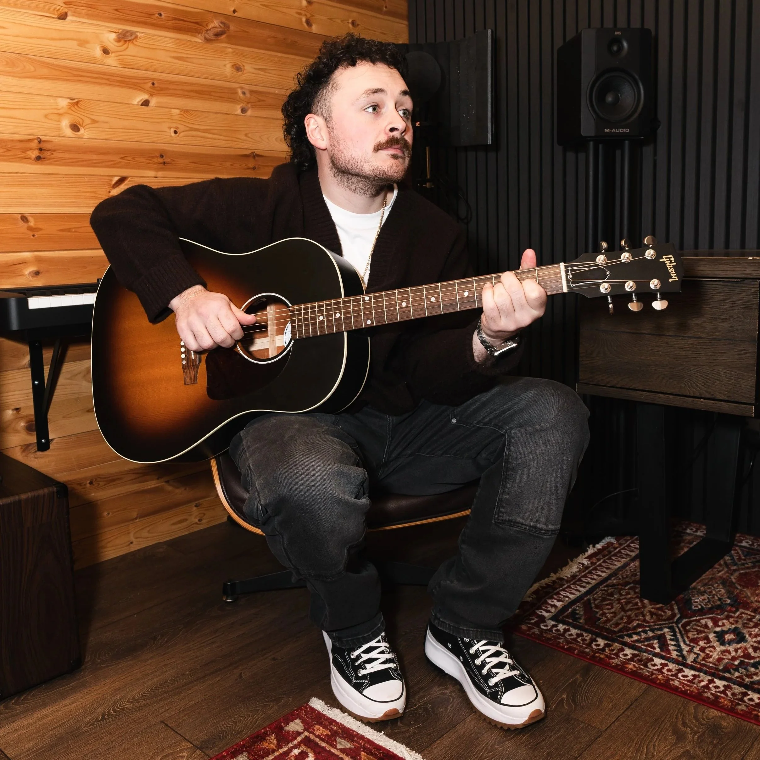 A man with curly dark hair and a beard, wearing a black cardigan, white t-shirt, dark jeans, and black-and-white sneakers, playing an acoustic guitar in a recording studio with wooden walls and audio equipment.
