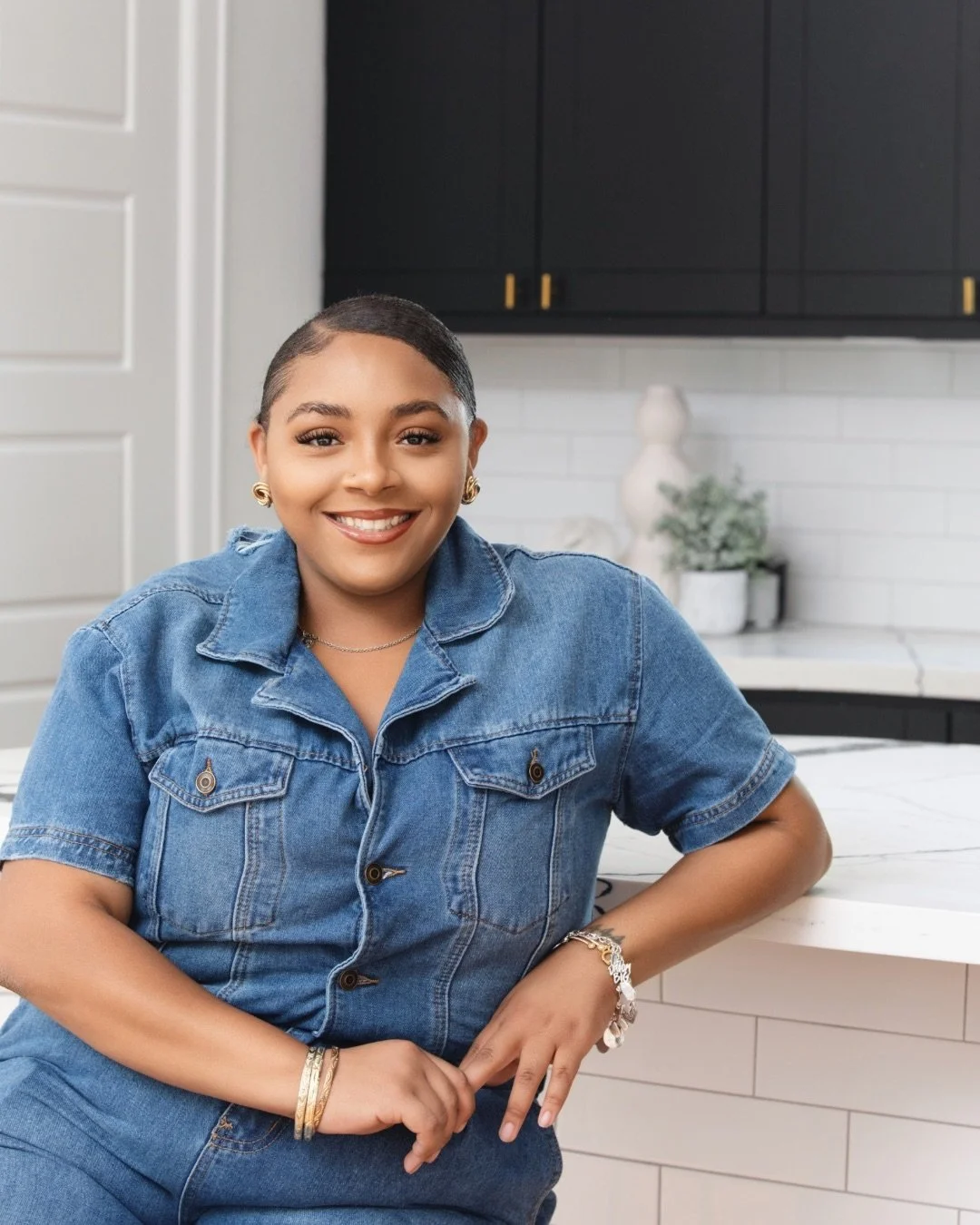 A woman with short hair and gold earrings, wearing a denim shirt and jewelry, smiling while sitting in a modern kitchen with white and black cabinets and a marble countertop.