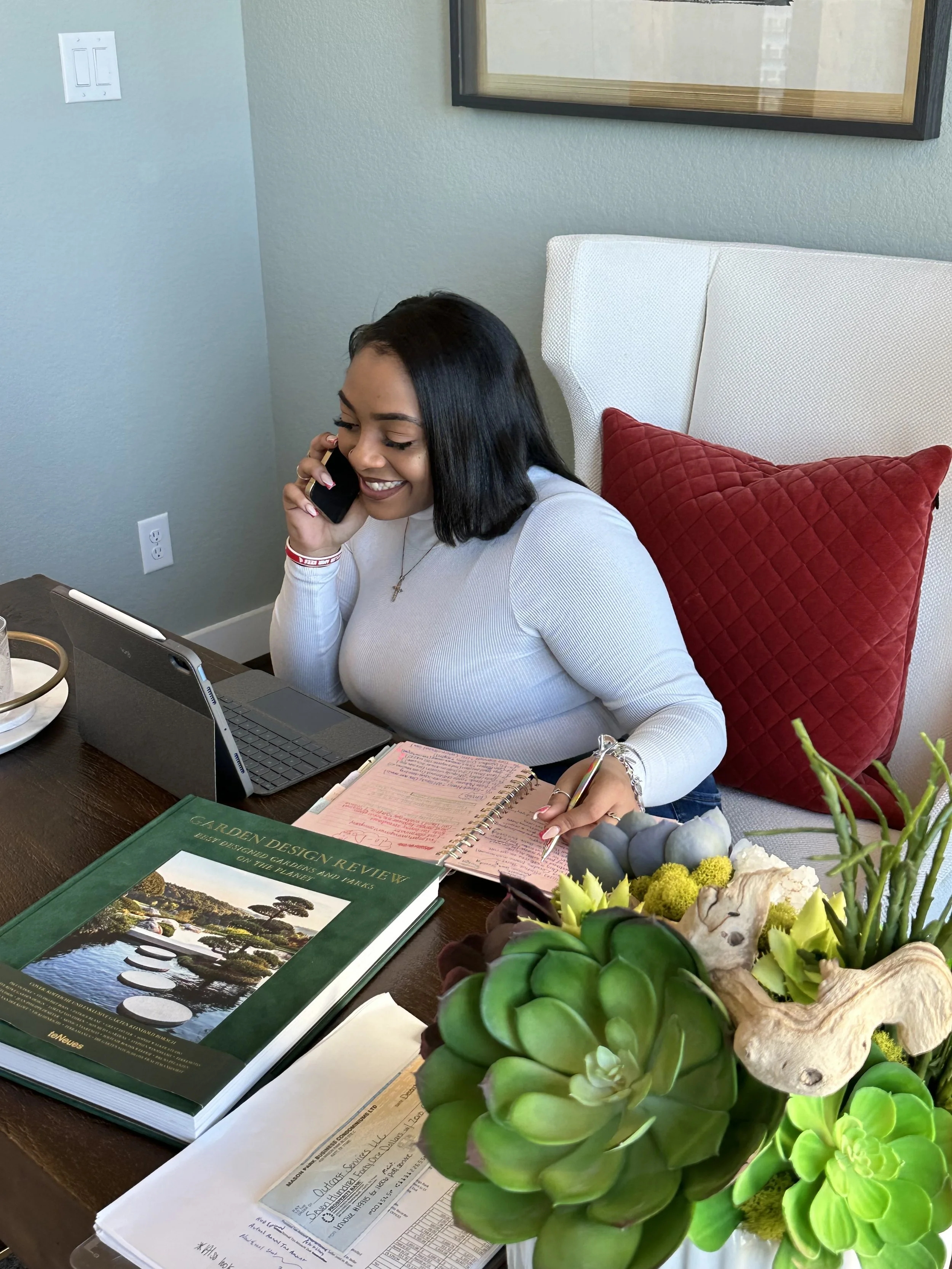 A woman sitting at a desk, talking on a phone, with a tablet, notes, and a large book titled 'Garden Design Review,' and a plant in the foreground.
