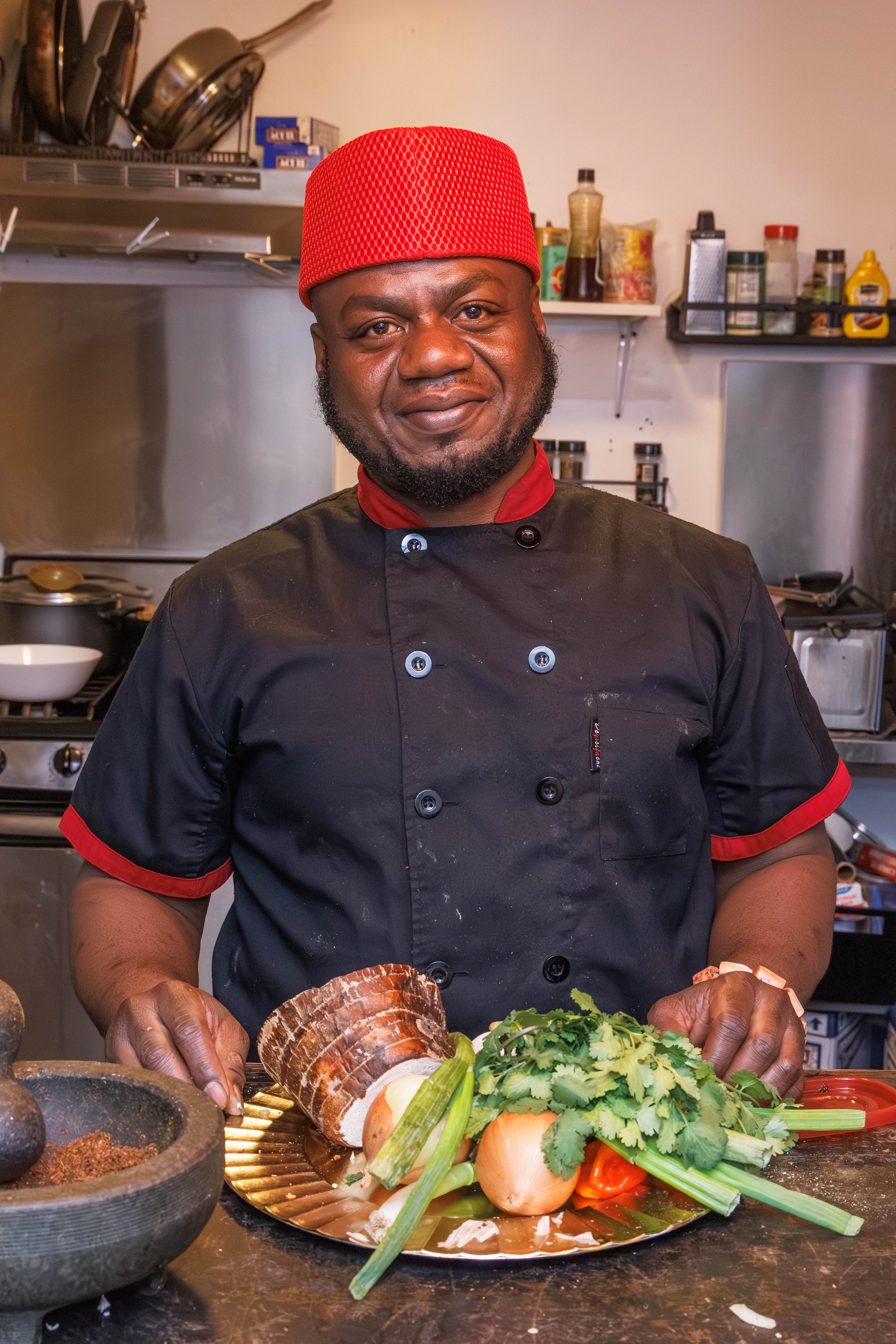 A chef standing in a kitchen with fresh vegetables on a plate, wearing a black chef's coat with red accents and a red hat.