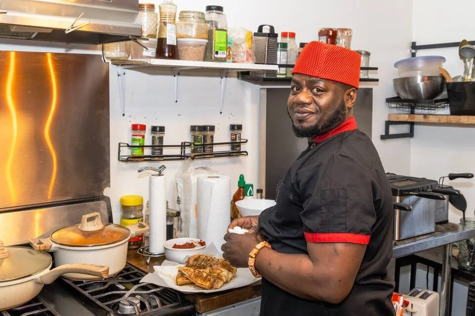 A man in a black chef's uniform with red trim and a red hat, standing in a professional kitchen, preparing food.