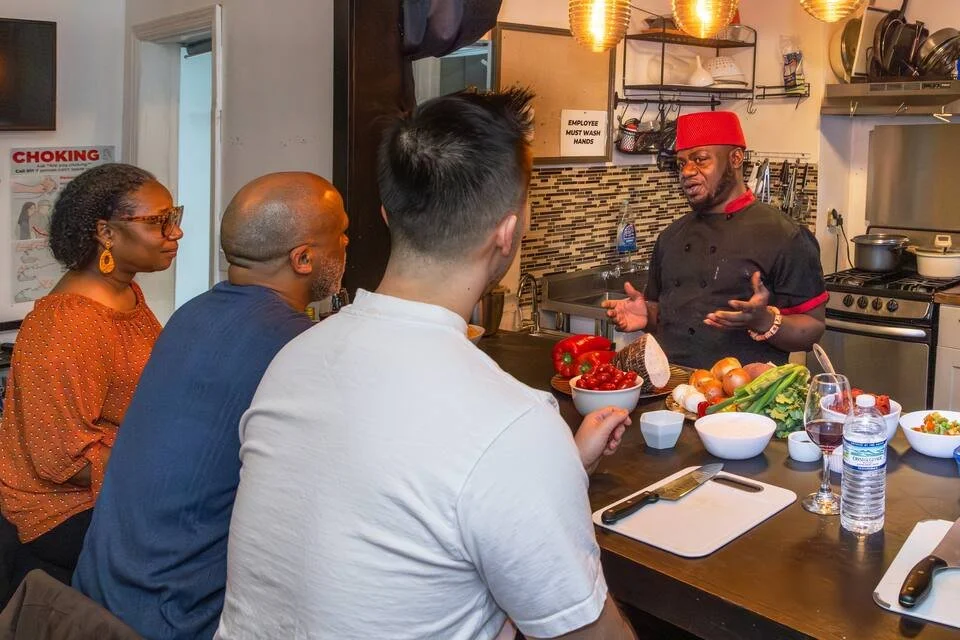 A chef in a black uniform with red accents is giving a cooking demonstration to three people seated at a kitchen counter. The counter has fresh vegetables, chopped ingredients, and bowls, with a kitchen background including hanging pots and cookware.