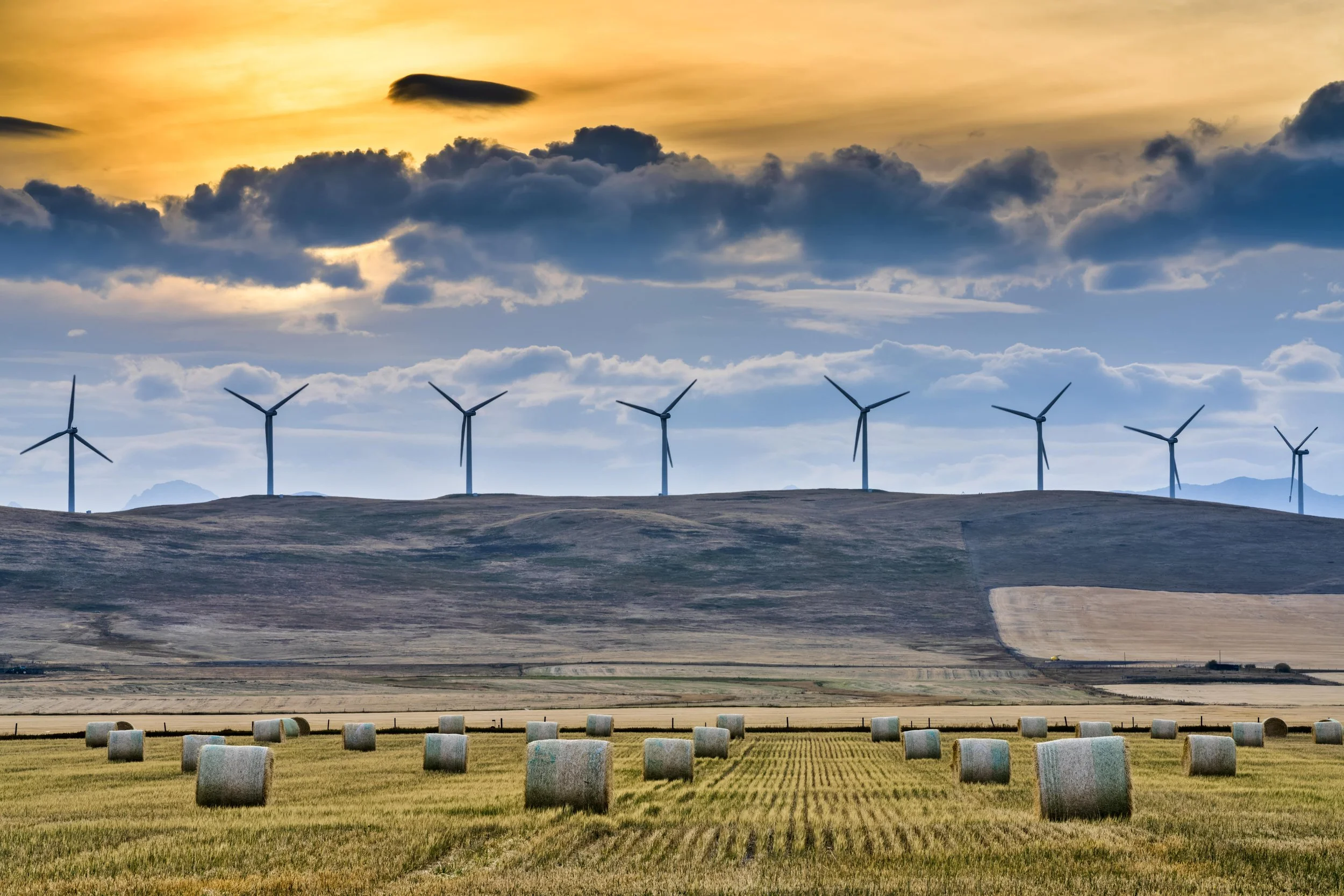 A landscape scene with wind turbines on a hill at sunset or sunrise, and a field with hay bales in the foreground.