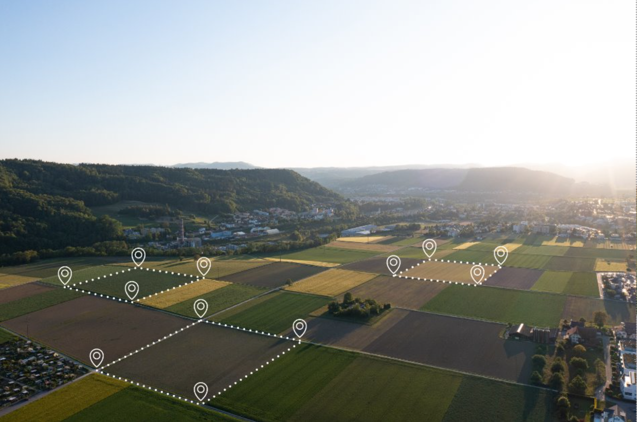 Aerial view of farmland with outlined plots marked by white pins, overlooking a town with mountains in the background during sunset.