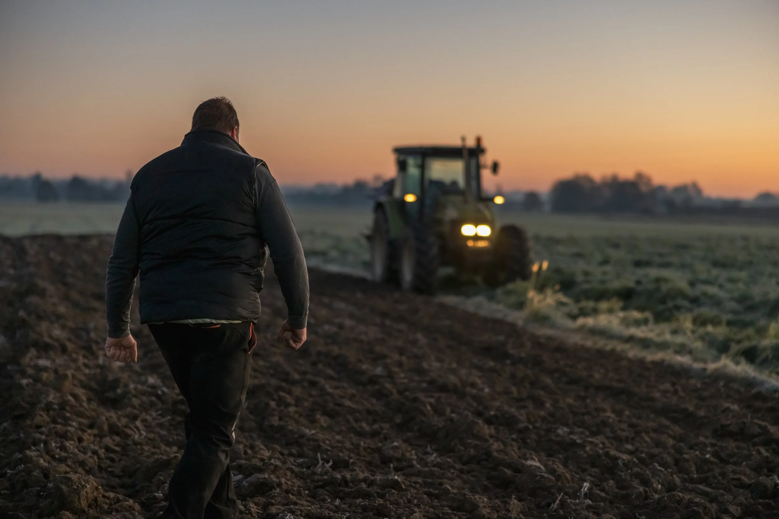 Man walks on farmland at sunset with tractor in the background.