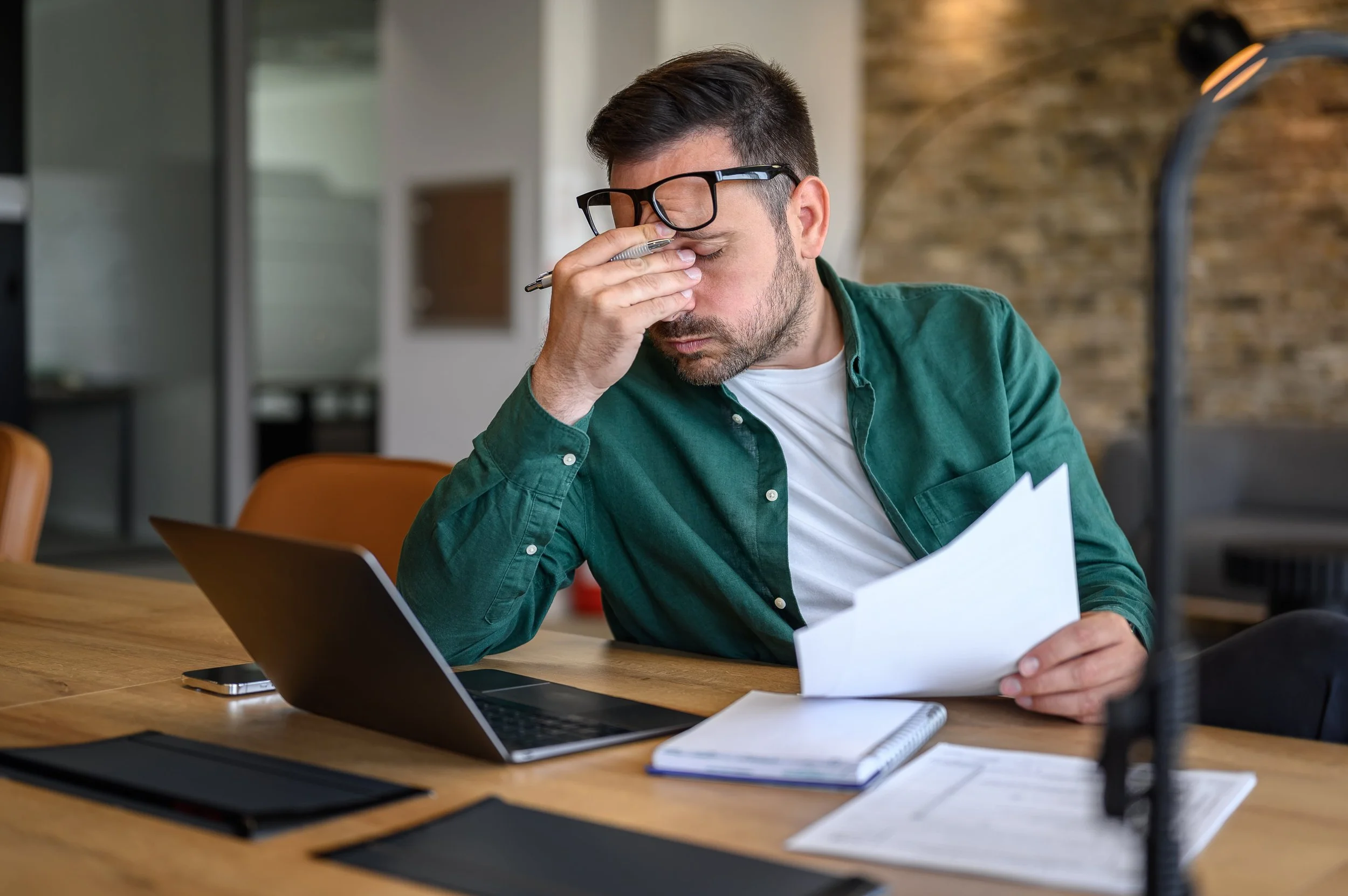 A stressed man in glasses, green shirt, and white t-shirt holding his forehead with one hand and a paper with the other, sitting at a desk with a laptop, notebook, and phone, in a modern office.