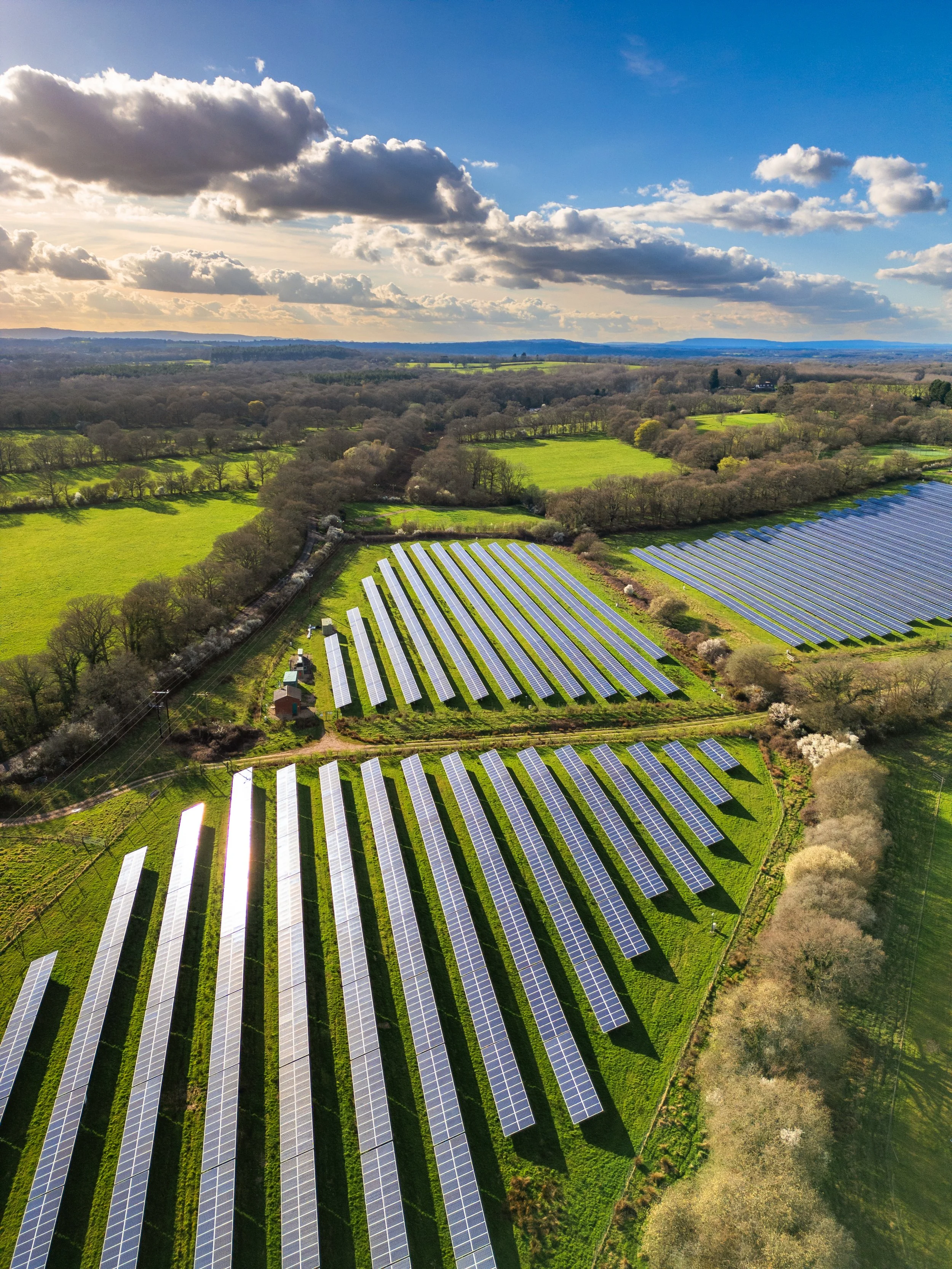 Aerial view of a solar farm with rows of solar panels on a green field, surrounded by trees and open countryside under a partly cloudy sky.