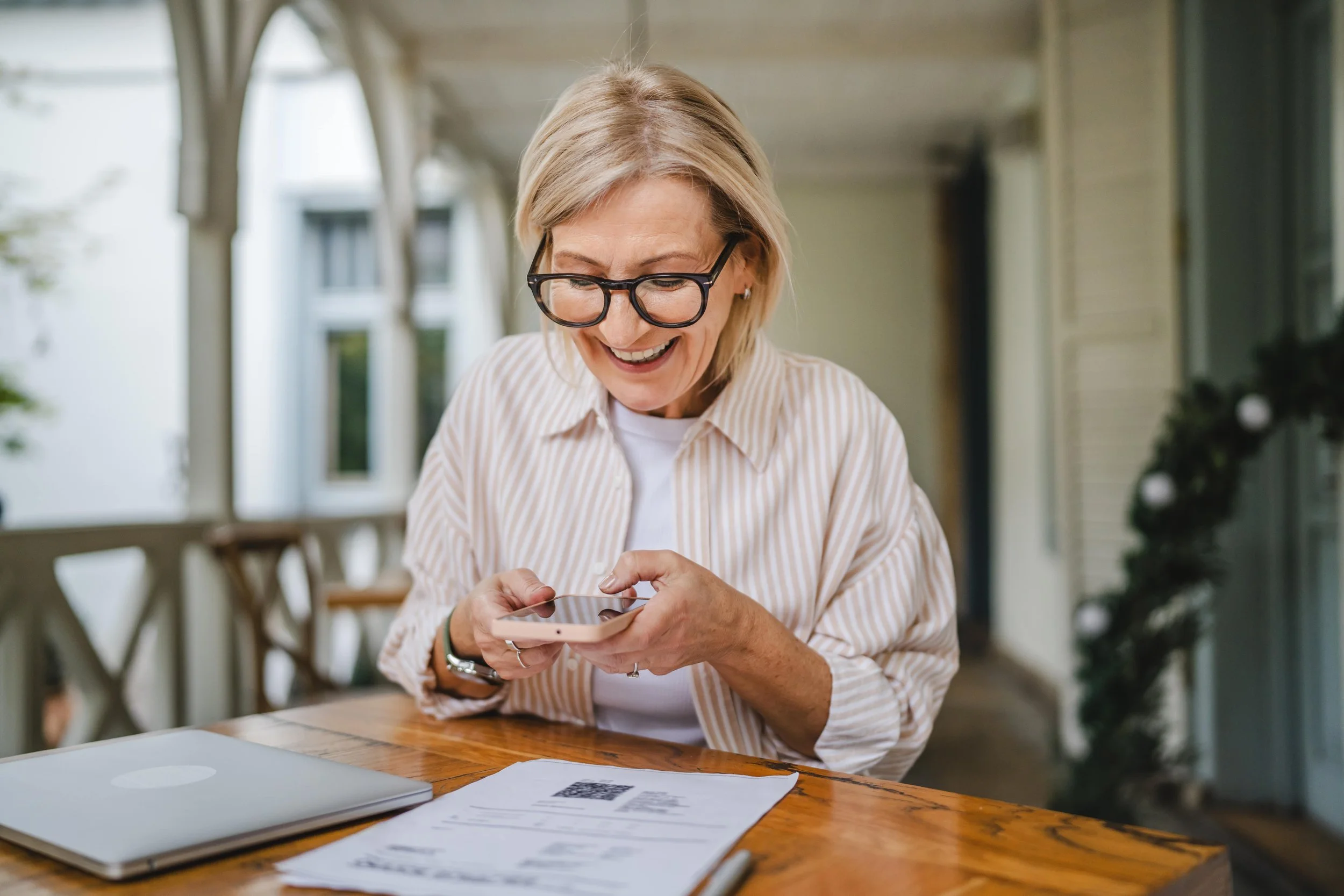 An elderly woman with glasses sitting at a wooden table, looking at her phone with a smile, with a closed laptop and papers on the table.