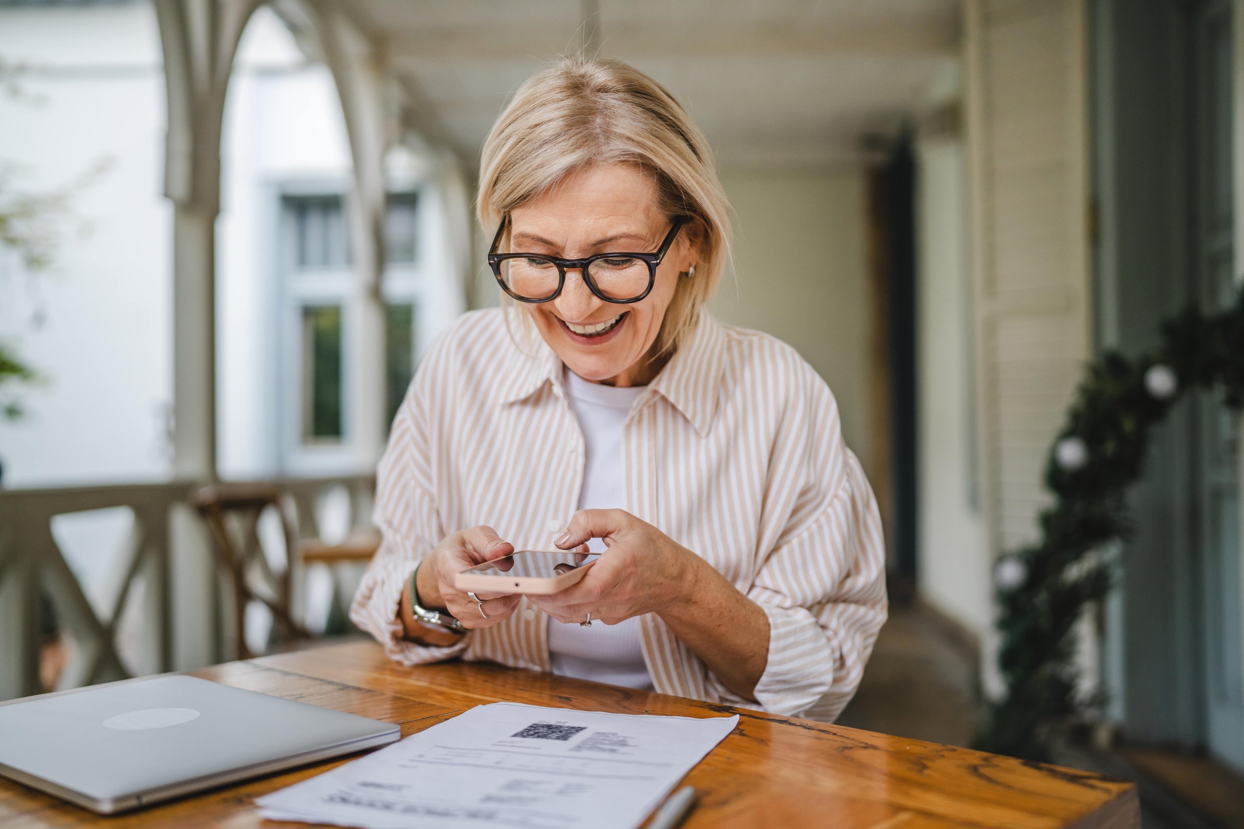 A woman with glasses smiling while looking at her phone, sitting at a wooden table with a laptop and papers, on a porch with holiday decorations.