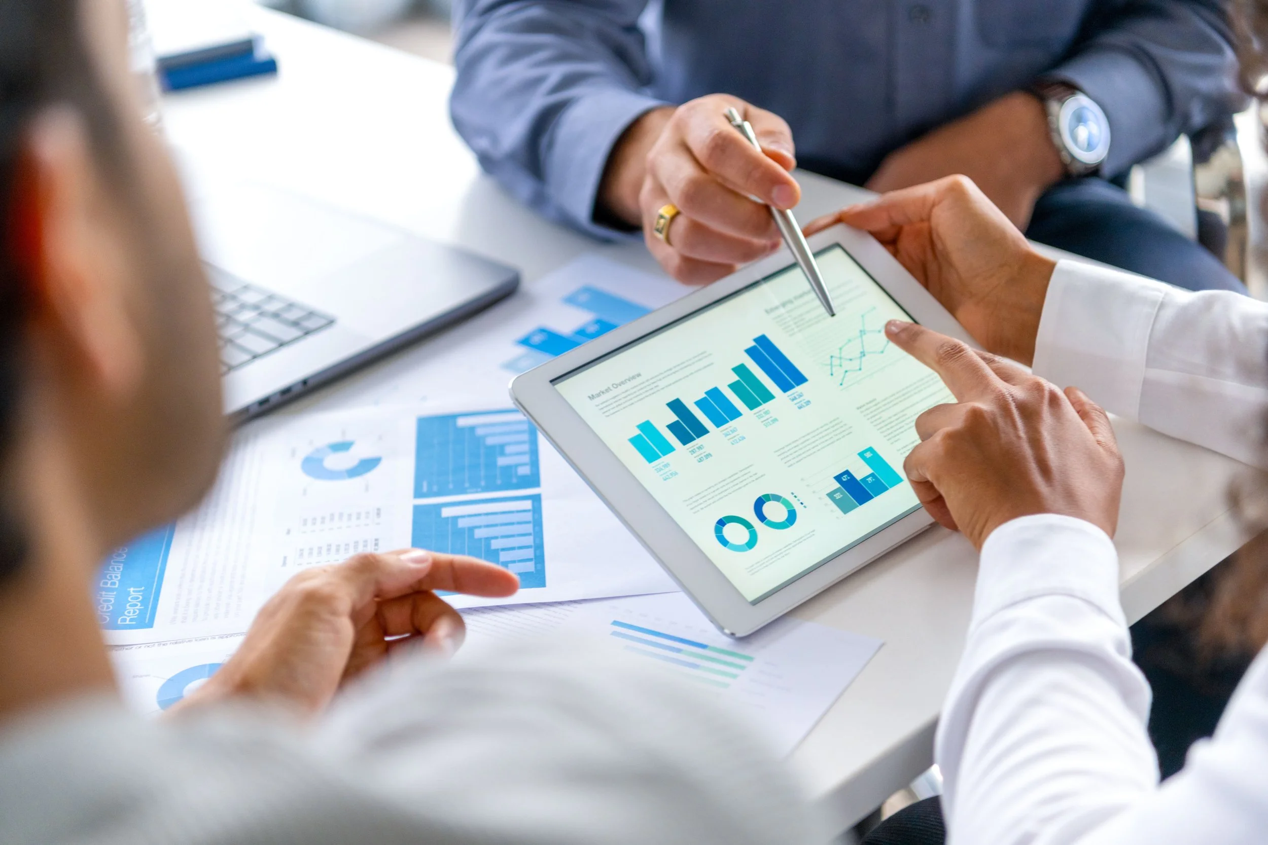People analyzing business data and charts on a digital tablet and printed reports with blue graphs at a meeting.