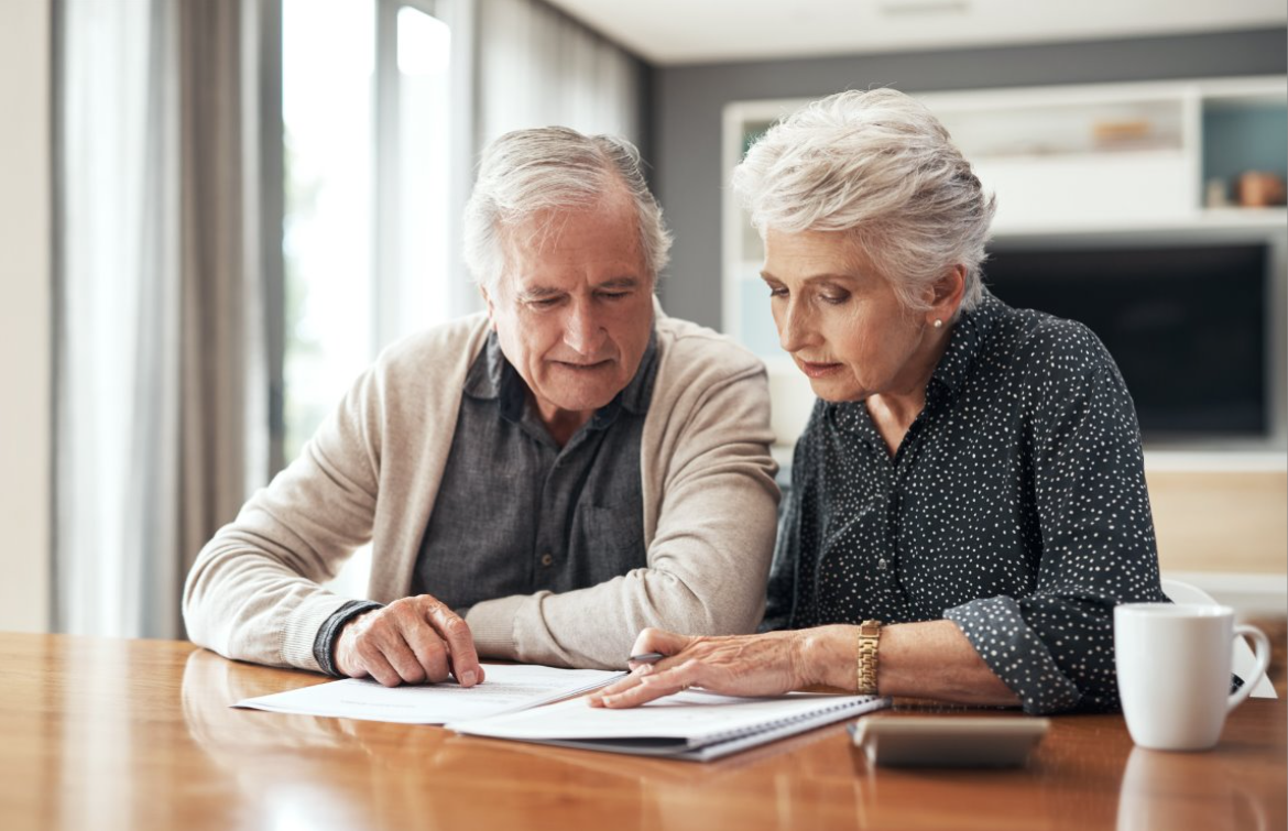 An elderly couple reviewing documents at a wooden table in a bright, modern kitchen with large windows.