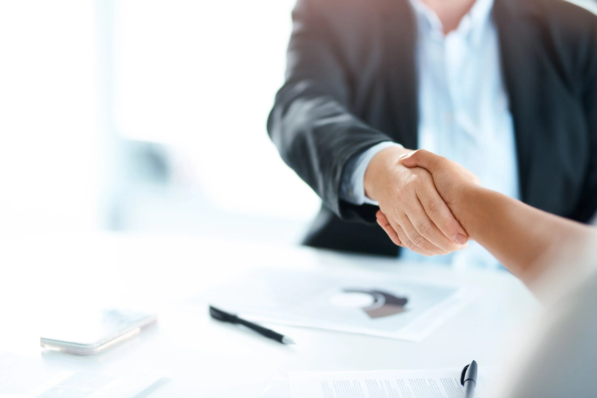 Close-up of two people shaking hands in a business setting with documents, a pen, and a smartphone on the table.