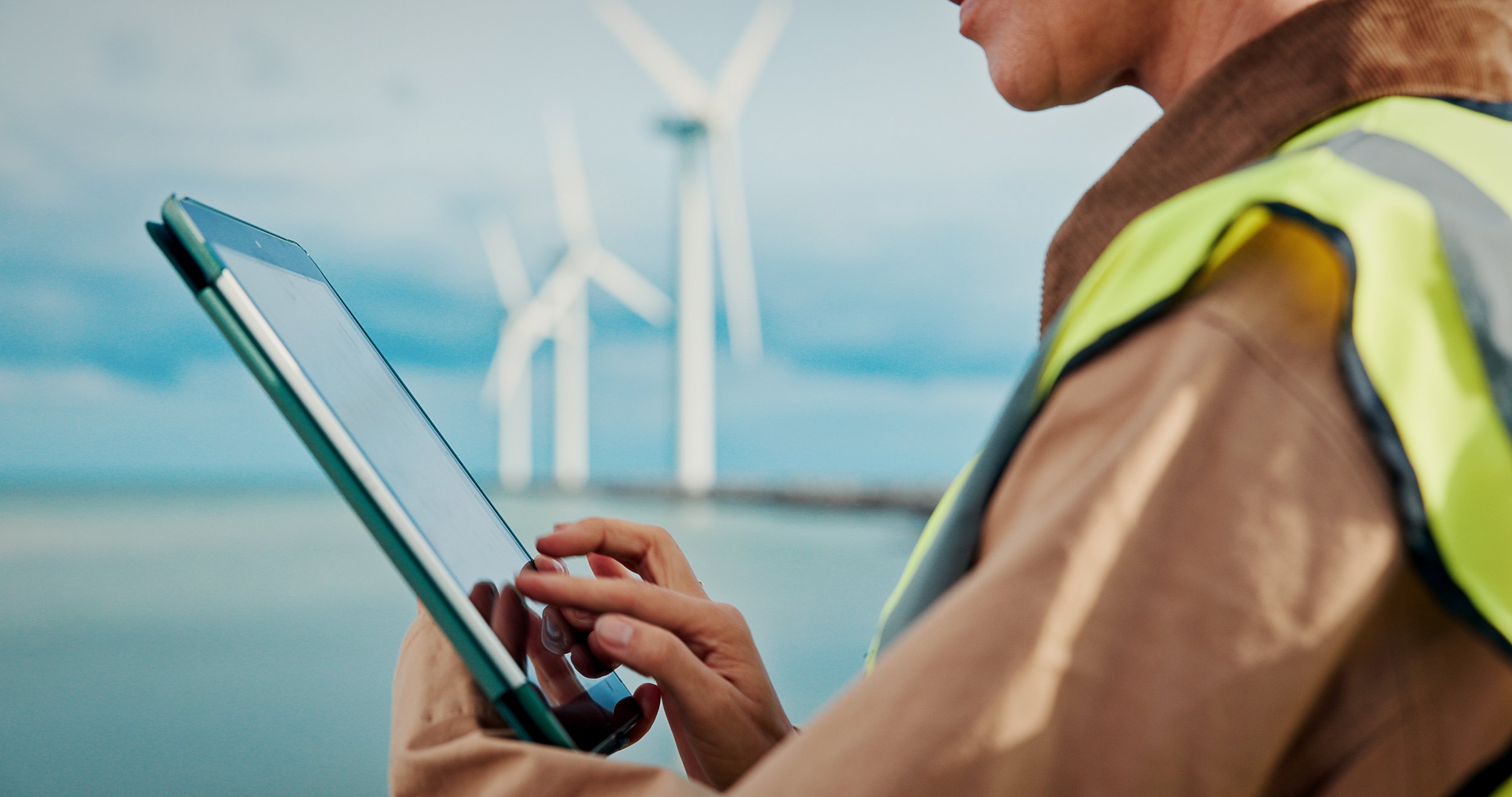 Person wearing a safety vest and brown jacket using a tablet in front of wind turbines near water.