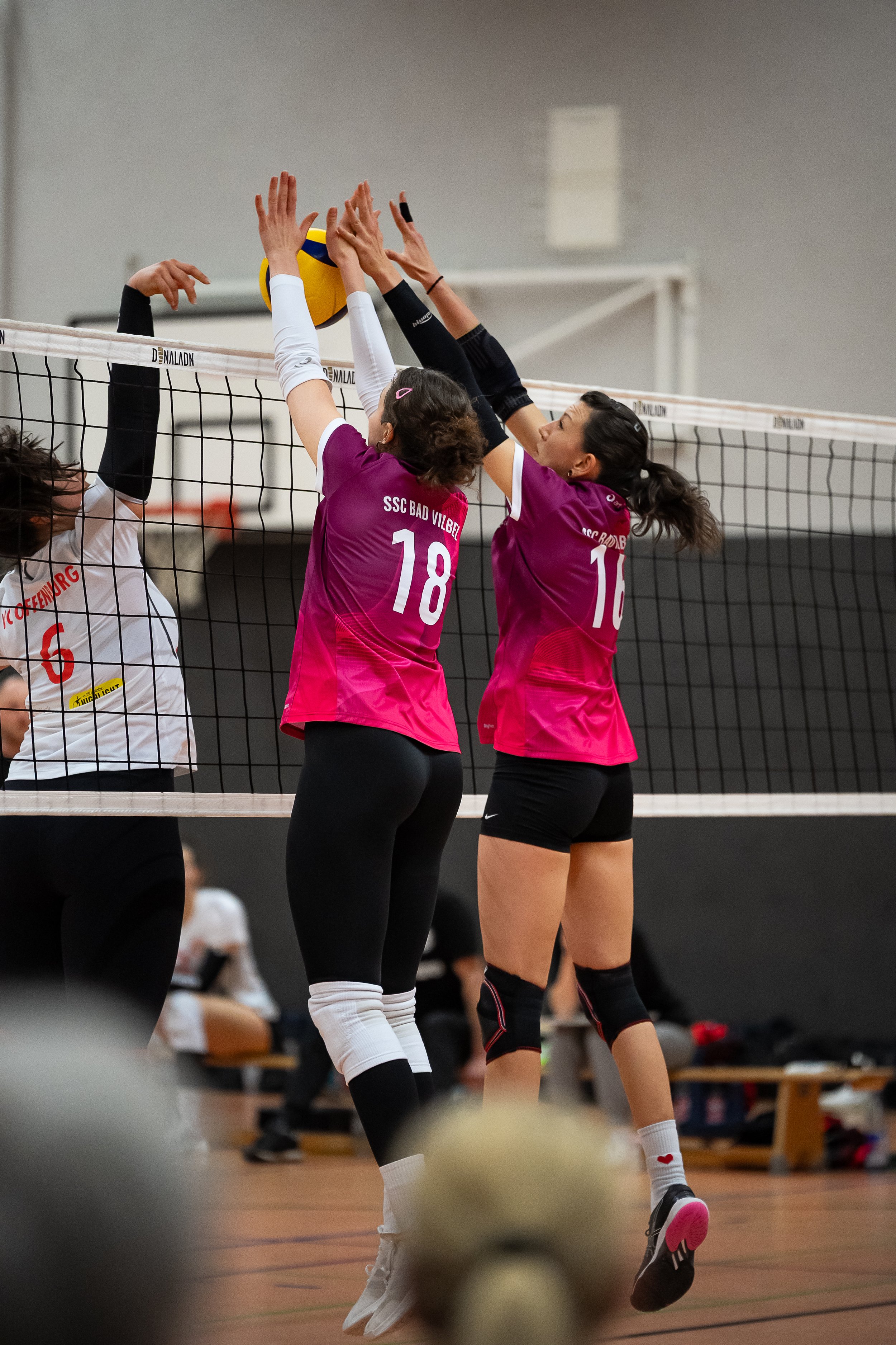 Zwei Volleyballspielerinnen in pinken Trikots blocken einen Ball am Netz, während eine Gegnerin einen Angriff ausführt.