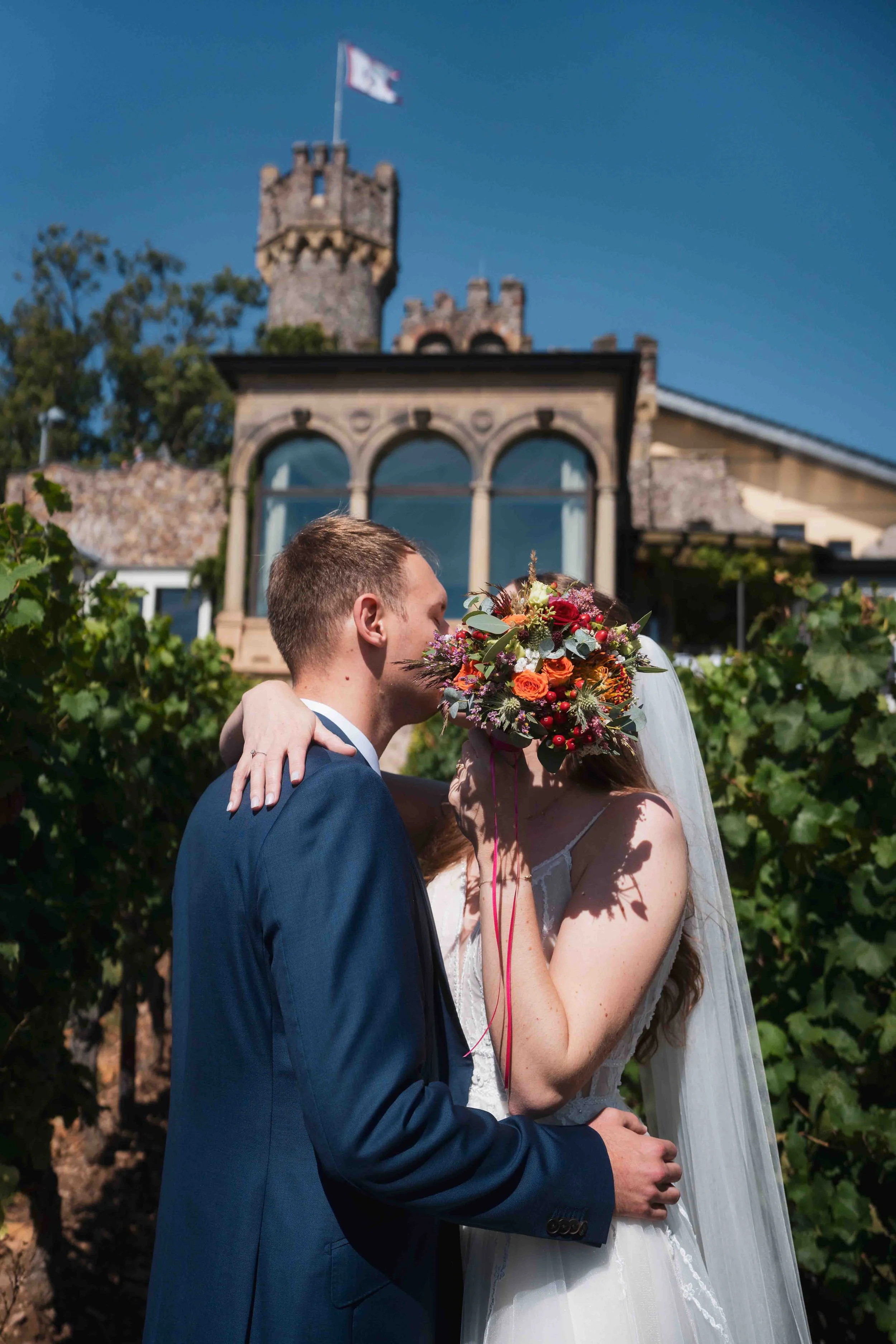 Ein Brautpaar bei einer Hochzeit im Freien, umrahmt von Weinreben, mit einer Burg im Hintergrund bei sonnigem Wetter.