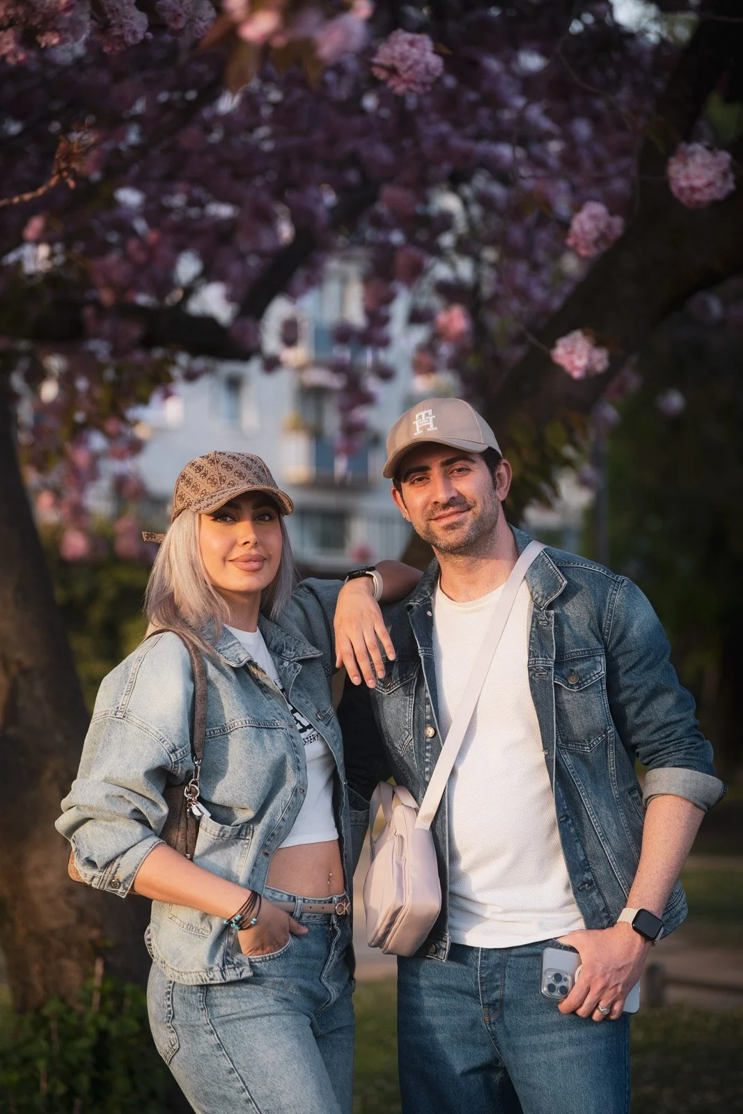 Ein Mann und eine Frau stehen vor einem blühenden Baum mit rosa Blumen, die einander umarmen und lächeln, in einem Park bei Sonnenuntergang.