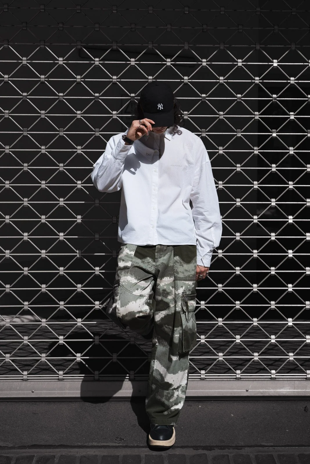 Person in Weißer Hemd, Camouflage-Hose und schwarzer New York Yankees Cap, lehnt gegen eine Metallgittertür.