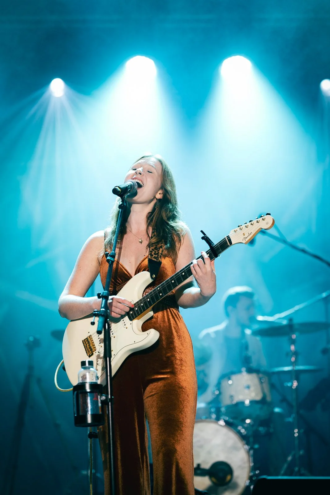 Eine Frau spielt Gitarre und singt bei einem Live-Konzert, im Hintergrund ein Musiker am Schlagzeug.