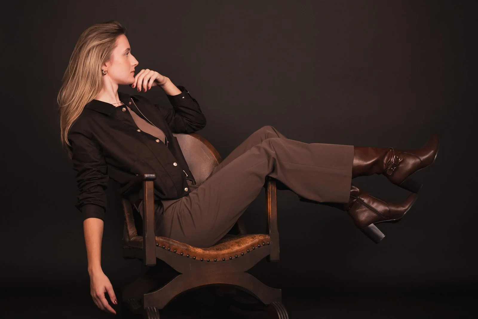 Frau sitzt seitlich auf einem antiken Holzstuhl, die Beine hochgelegt und trägt braune Stiefel, schwarze Jacke und braune Hosen vor einem schwarzen Hintergrund.