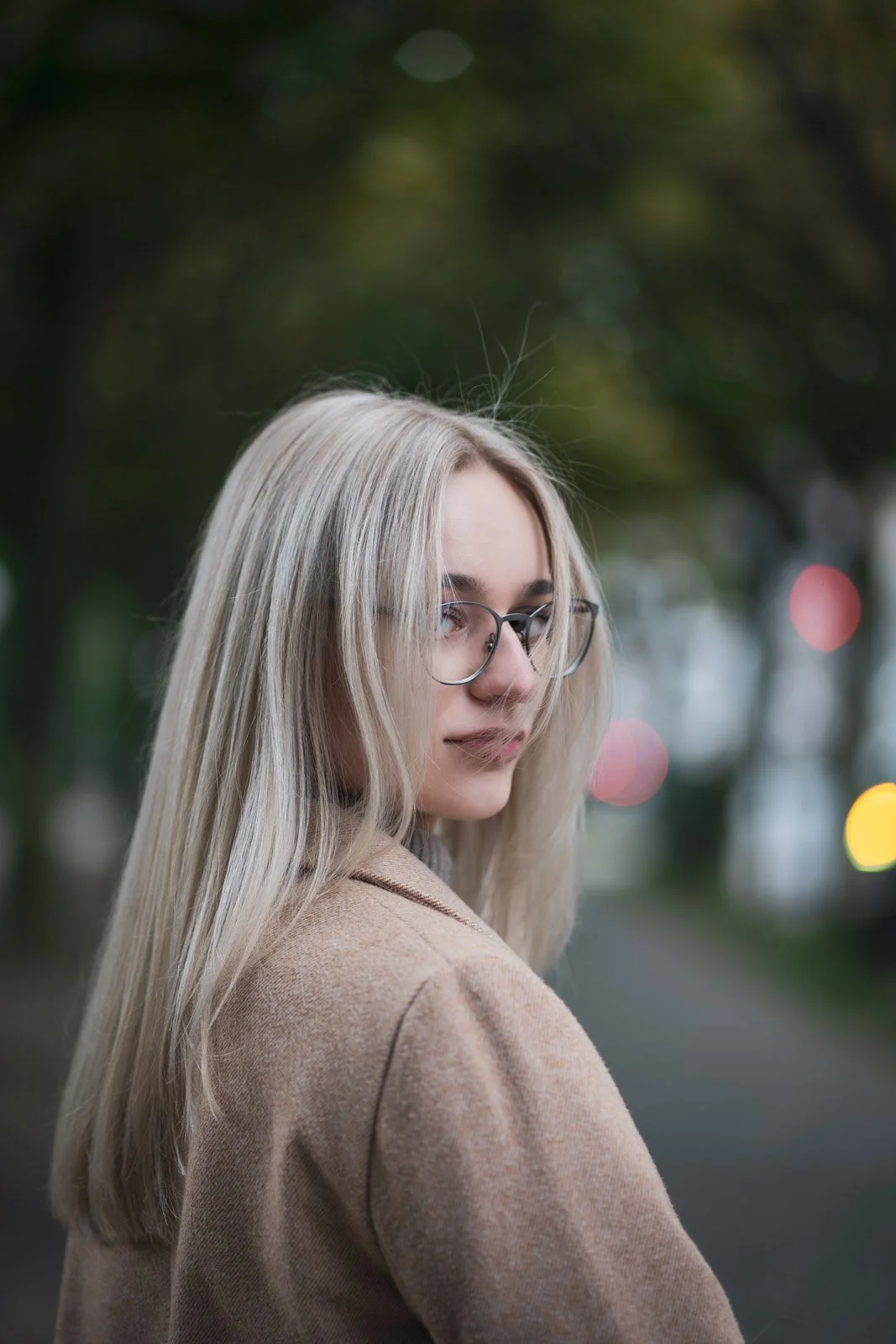 Junge Frau mit langem blondem Haar und Brille, trägt einen beigen Mantel, steht im Freien, im Hintergrund unscharfe Lichter und Bäume.