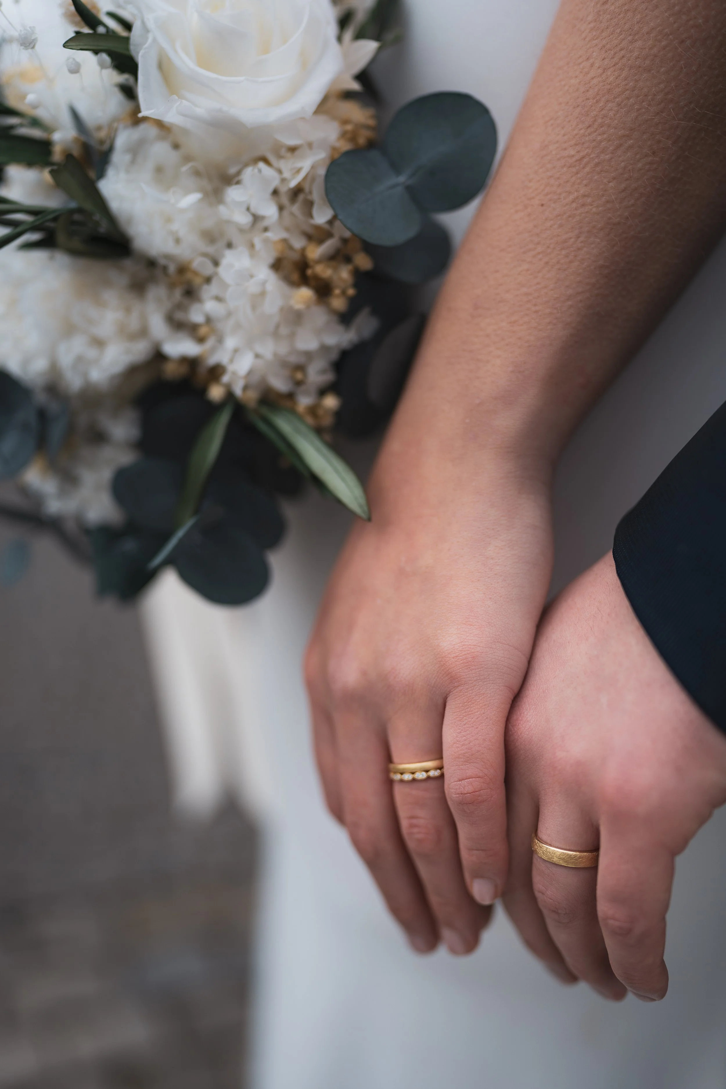 Nahaufnahme von zwei Händen, die sich halten, mit Eheringen, neben einem Blumenstrauß aus weißen Blumen und grünen Blättern.