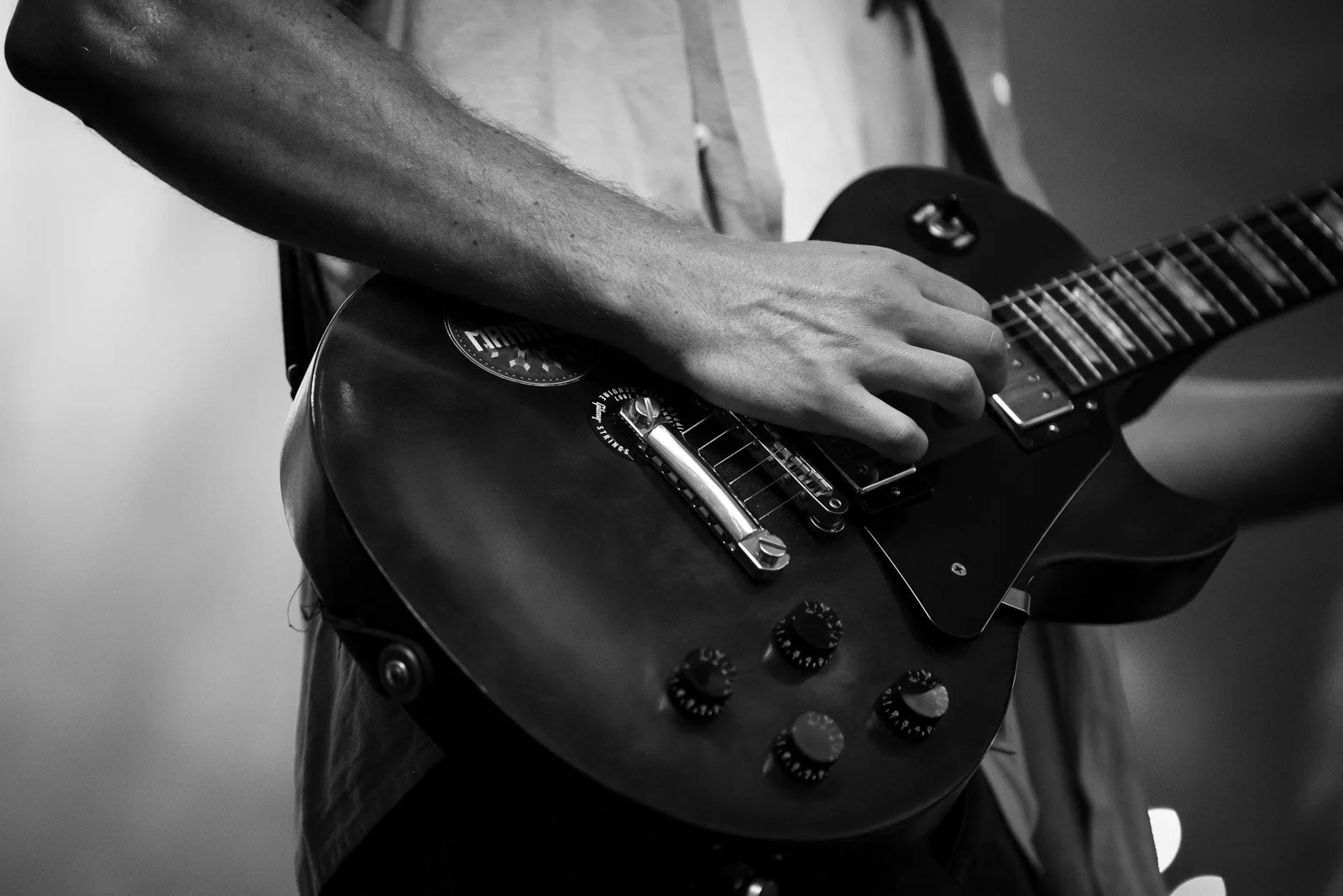Nahaufnahme einer Person, die eine elektrische Gitarre spielt, in schwarz-weiß. Die Person greift die Saiten der Gitarre, die Knöpfe und den Tonabnehmer sind sichtbar.