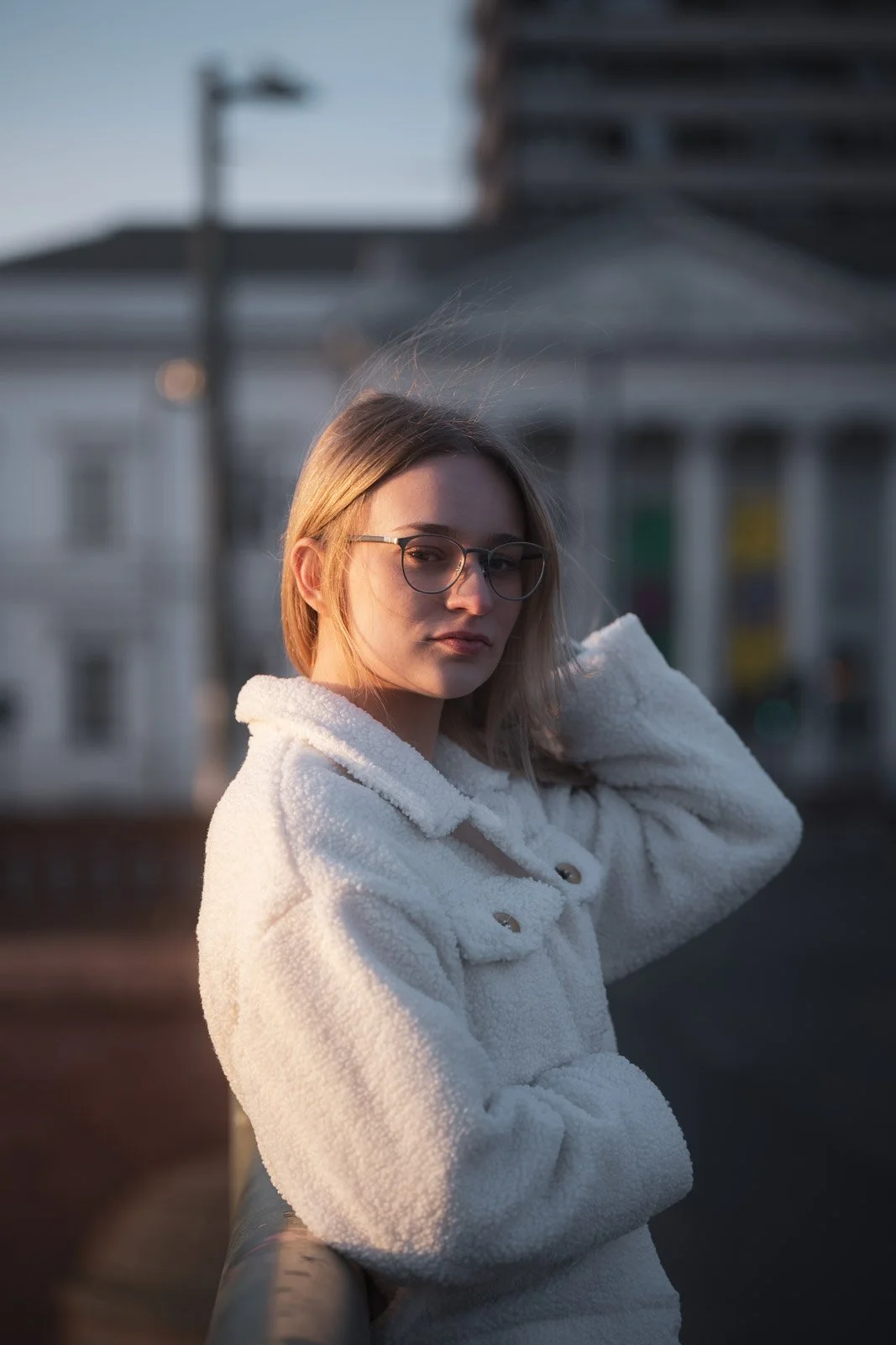 Junge Frau mit Brille trägt einen weißen Pullover und steht draußen bei Sonnenuntergang in einer Stadt.