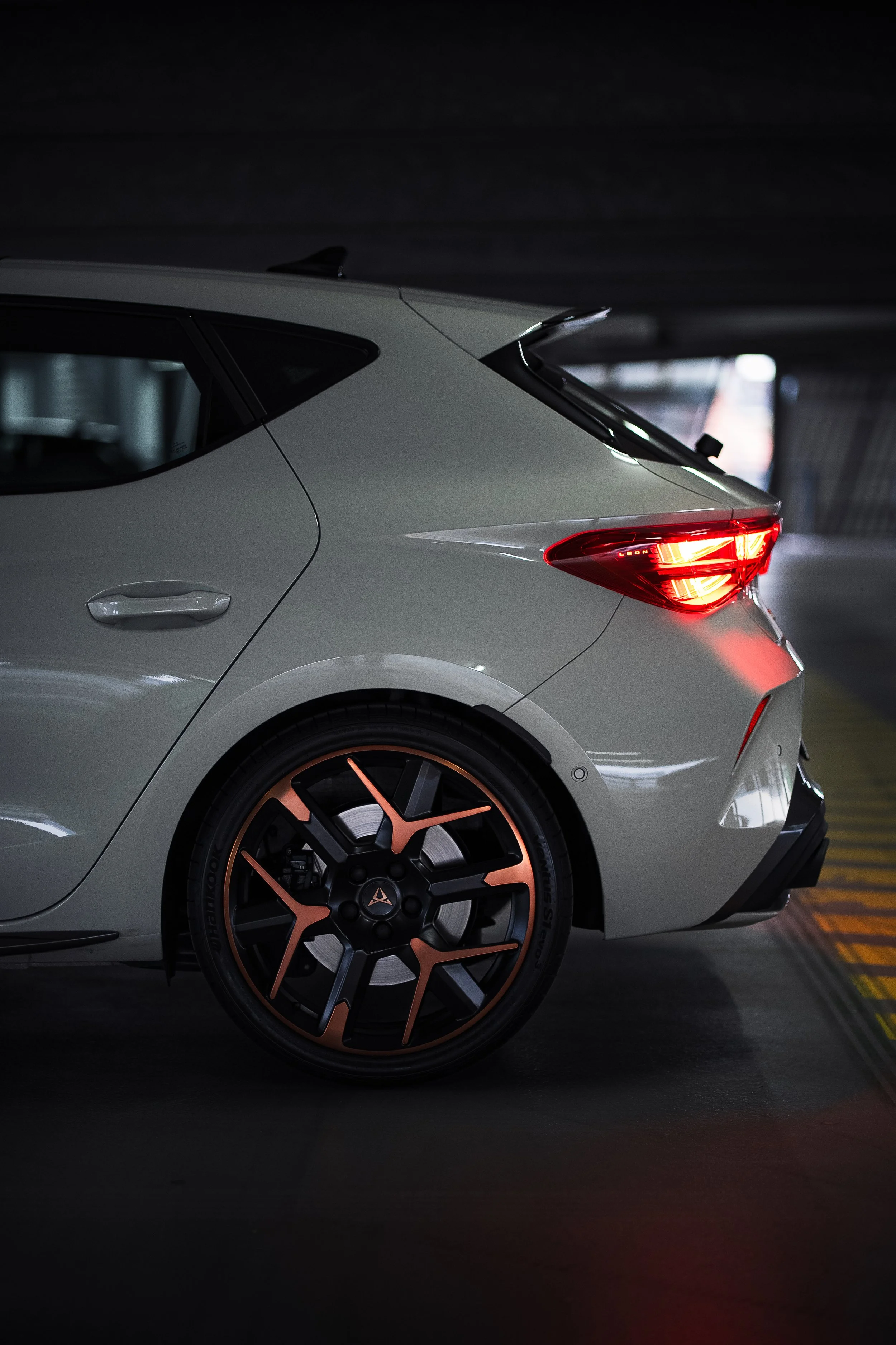 Nahaufnahme der hinteren Seite eines weißen Autos, das in einer Tiefgarage parkt. Das Auto hat einzigartige schwarze und kupferfarbene Felgen und leuchtende Rücklichter.
