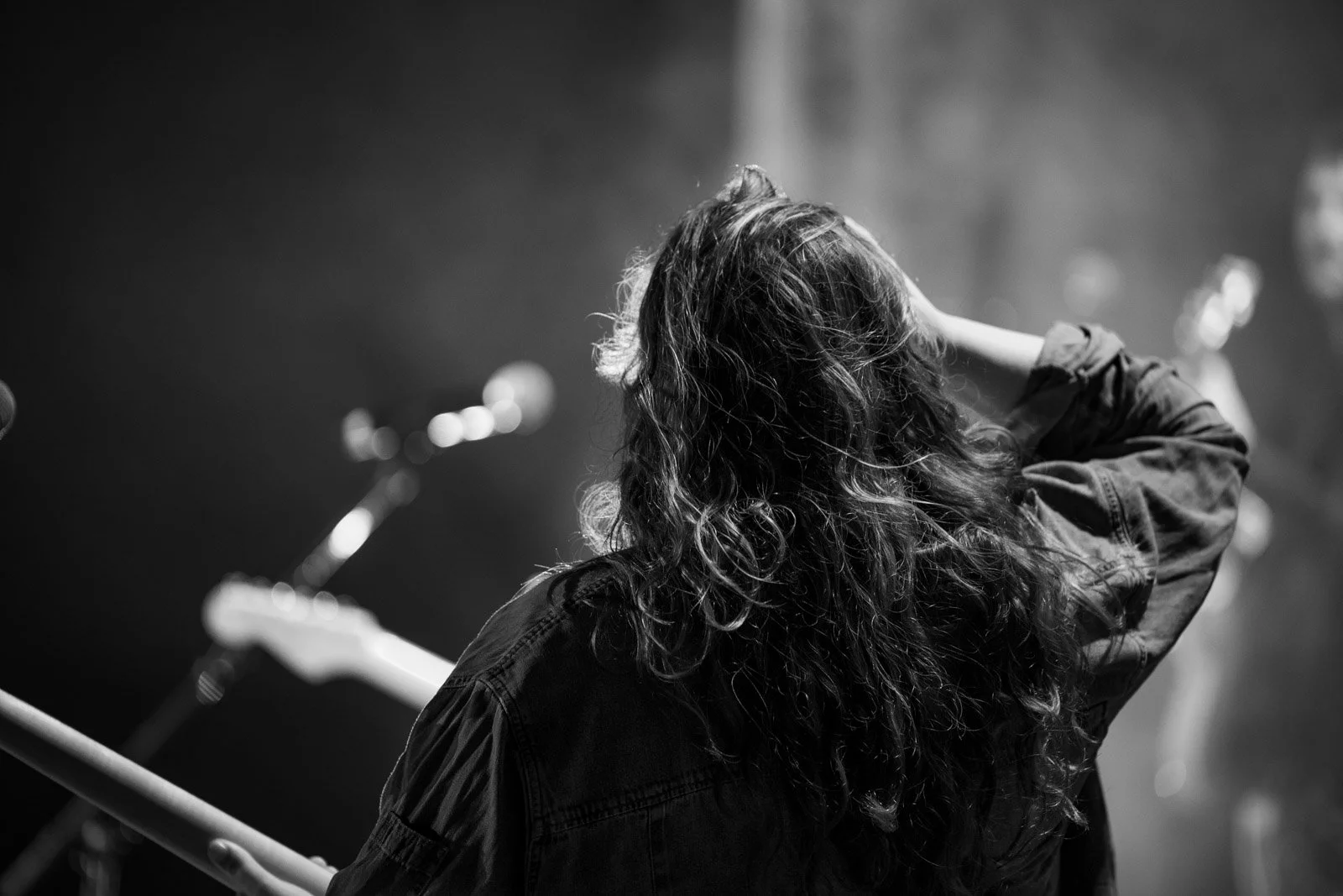 Schwarz-Weiß-Foto einer Frau mit langen, gewellten Haaren, die auf einer Bühne steht, möglicherweise bei einem Musik- oder Theaterauftritt, mit Mikrofonen im Hintergrund.