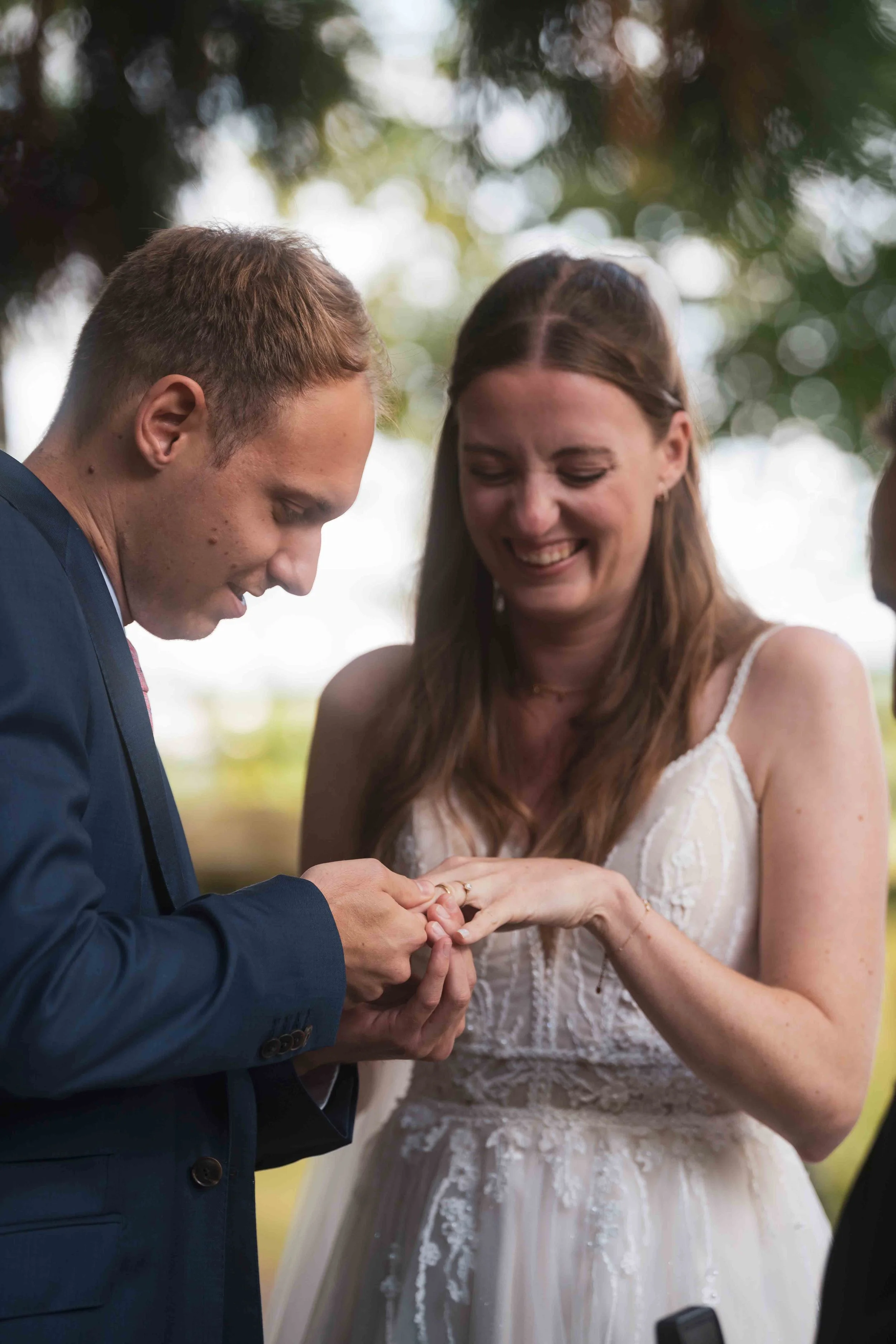 Ein Mann in Anzug steckt einem lächelnden Frau ein Ring an den Finger bei einer Hochzeitszeremonie im Freien.