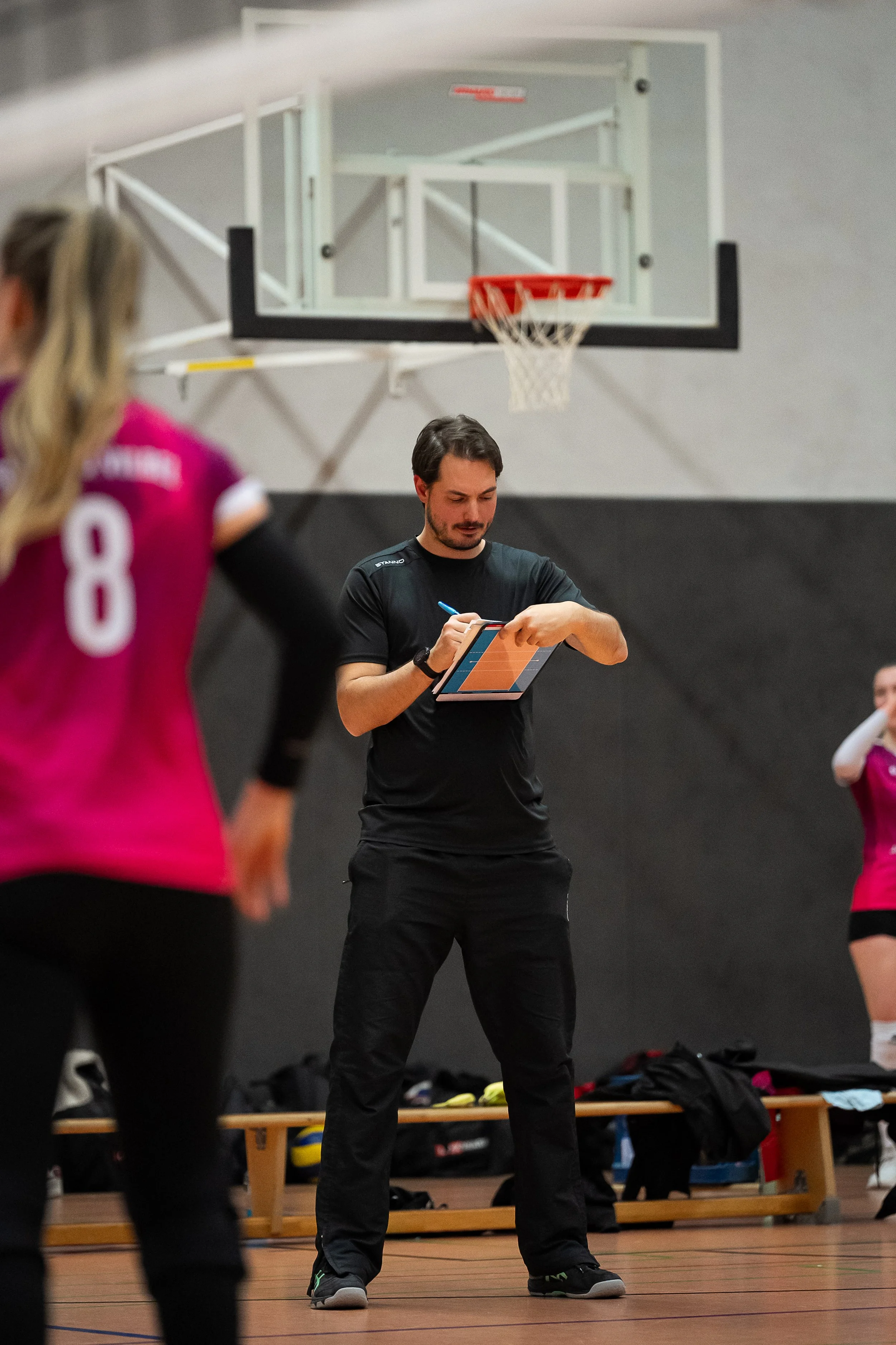 Ein männlicher Trainer auf einem Volleyballfeld schreibt auf ein Whiteboard, während zwei Spielerinnen in pinken Trikots im Hintergrund zuschauen.