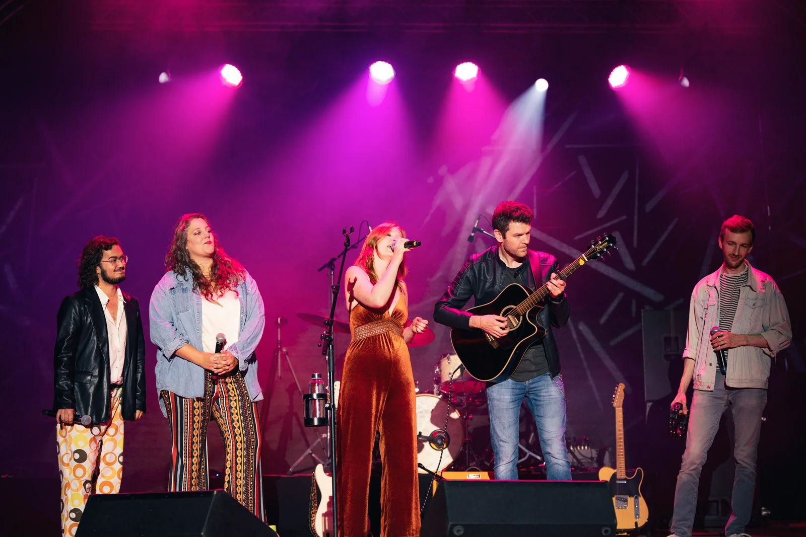 Musiker auf einer Bühne bei einem Konzert, ein Schlagzeug, eine Gitarre und fünf Sänger, bunte Beleuchtung.