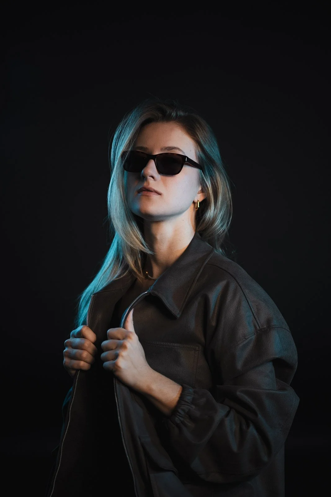 Junge Frau trägt eine schwarze Lederjacke und Sonnenbrille, posiert vor schwarzem Hintergrund.