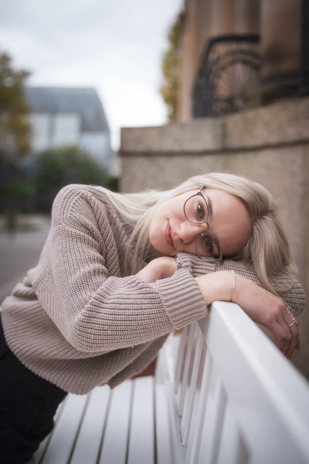 Junge Frau mit Brille liegt entspannt auf einer weißen Parkbank im Freien, trägt bequemen Pullover, während sie in die Kamera lächelt.