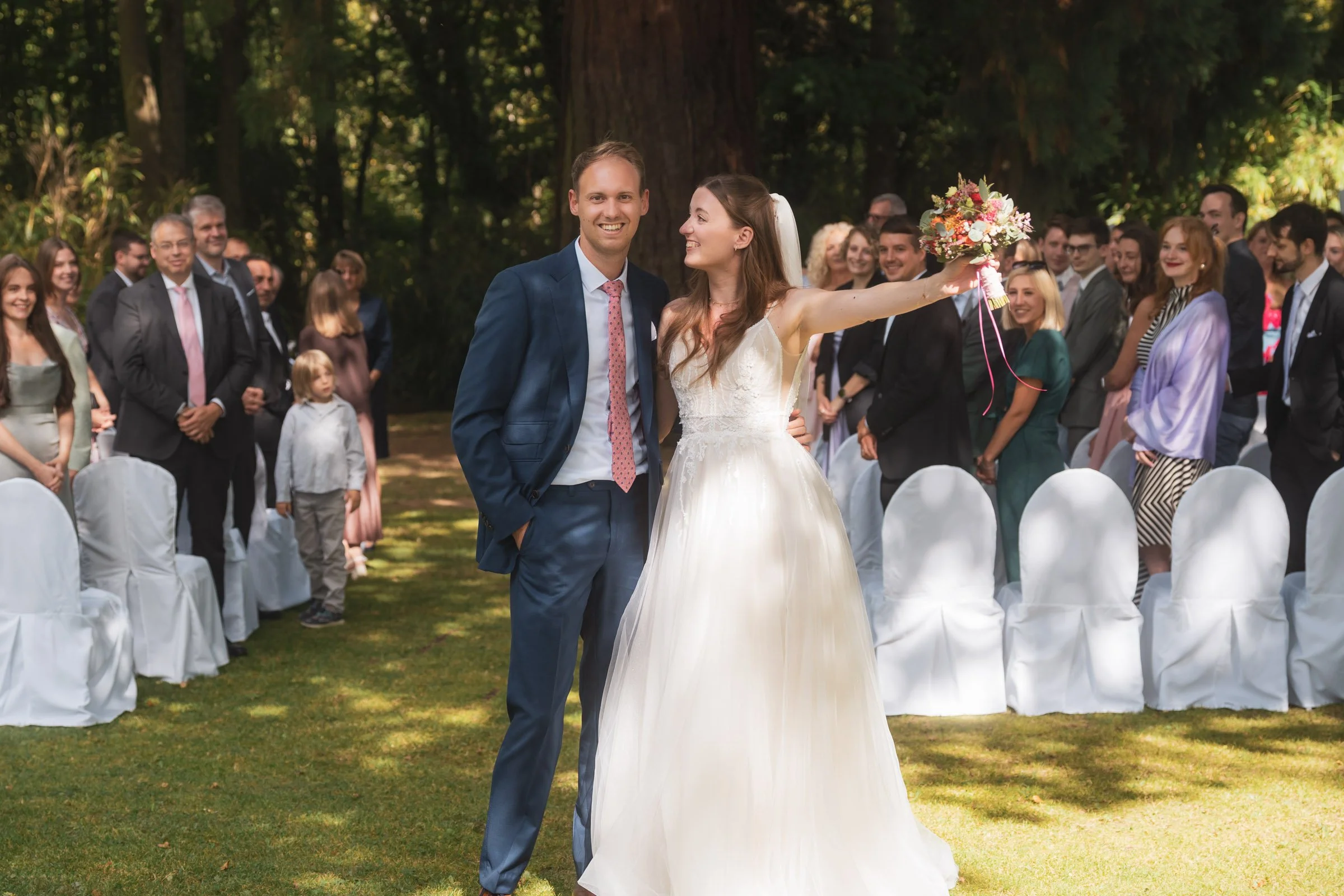 Braut wirft Blumenstrauß bei einer Hochzeit im Freien, umgeben von Gästen.