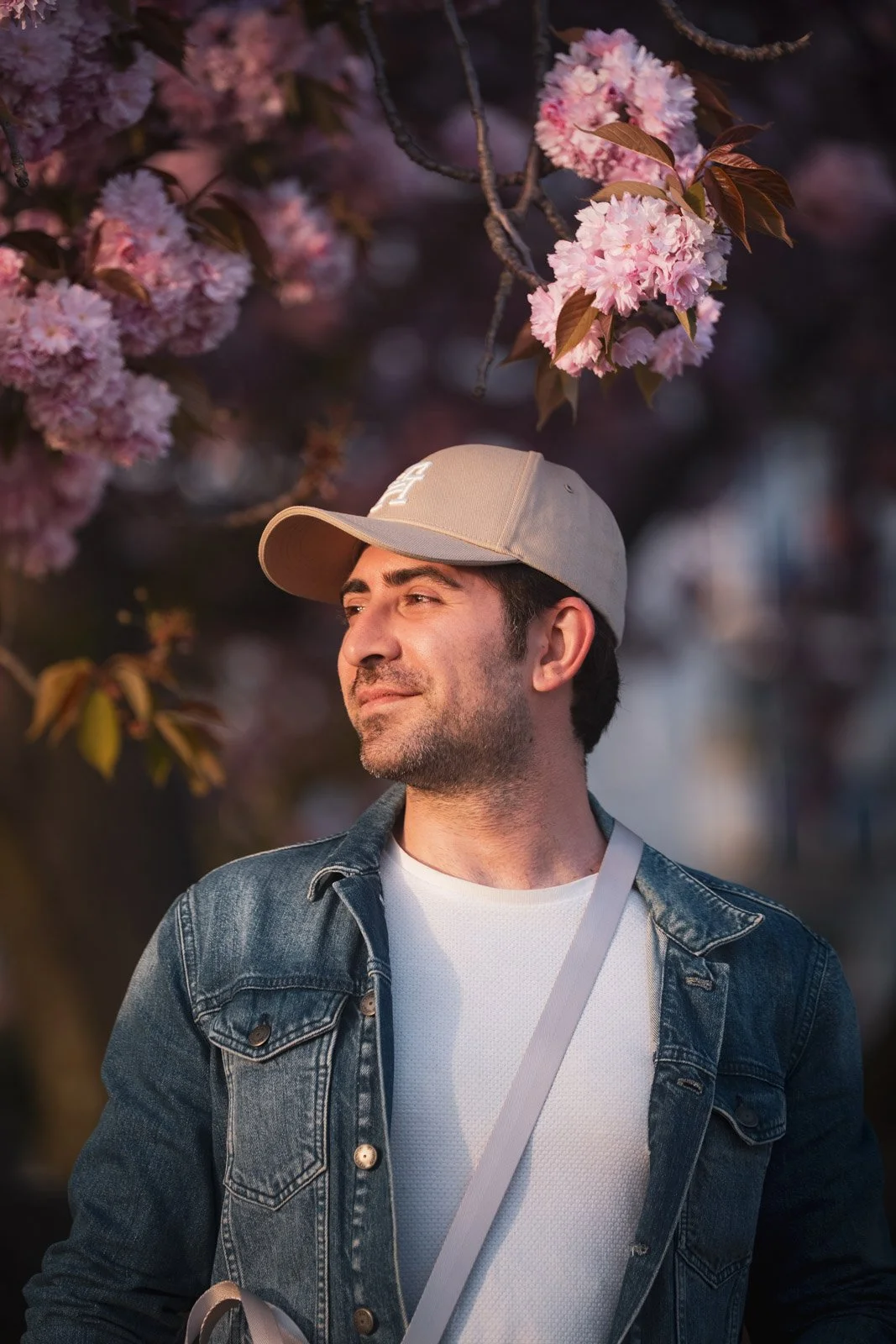 Ein Mann trägt eine weiße Mütze, einen dunkelblauen Jeansjacke und ein weißes T-Shirt, steht vor einem blühenden Kirschbaum und schaut nach rechts, bei Sonnenuntergang.