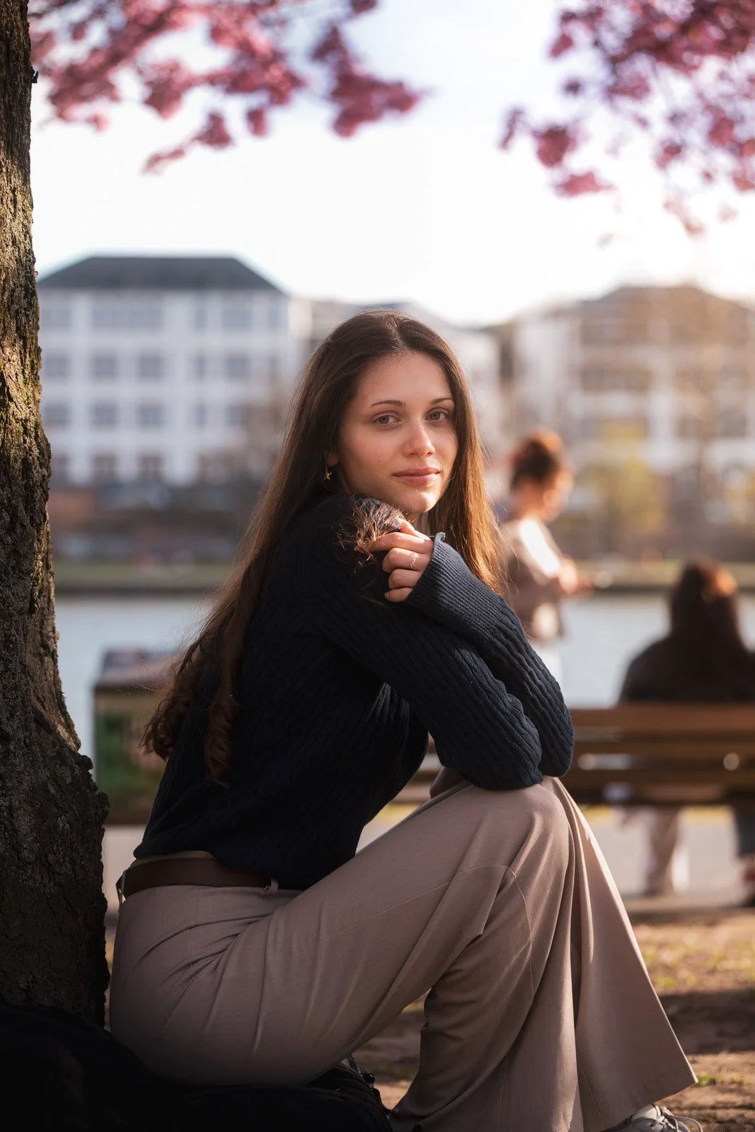 Junge Frau sitzt am Flussufer unter einem Baum, blickt in die Kamera, mit Gebäuden im Hintergrund, im Freien bei sonntäglichem Spaziergang, im Herbst.