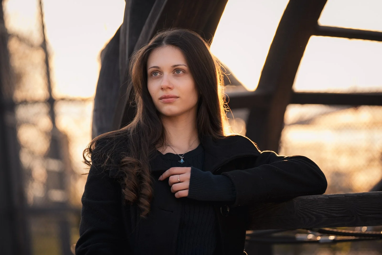 Junge Frau mit dunklem Haar und schwarze Kleidung steht an einer Holzkonstruktion bei Sonnenuntergang.