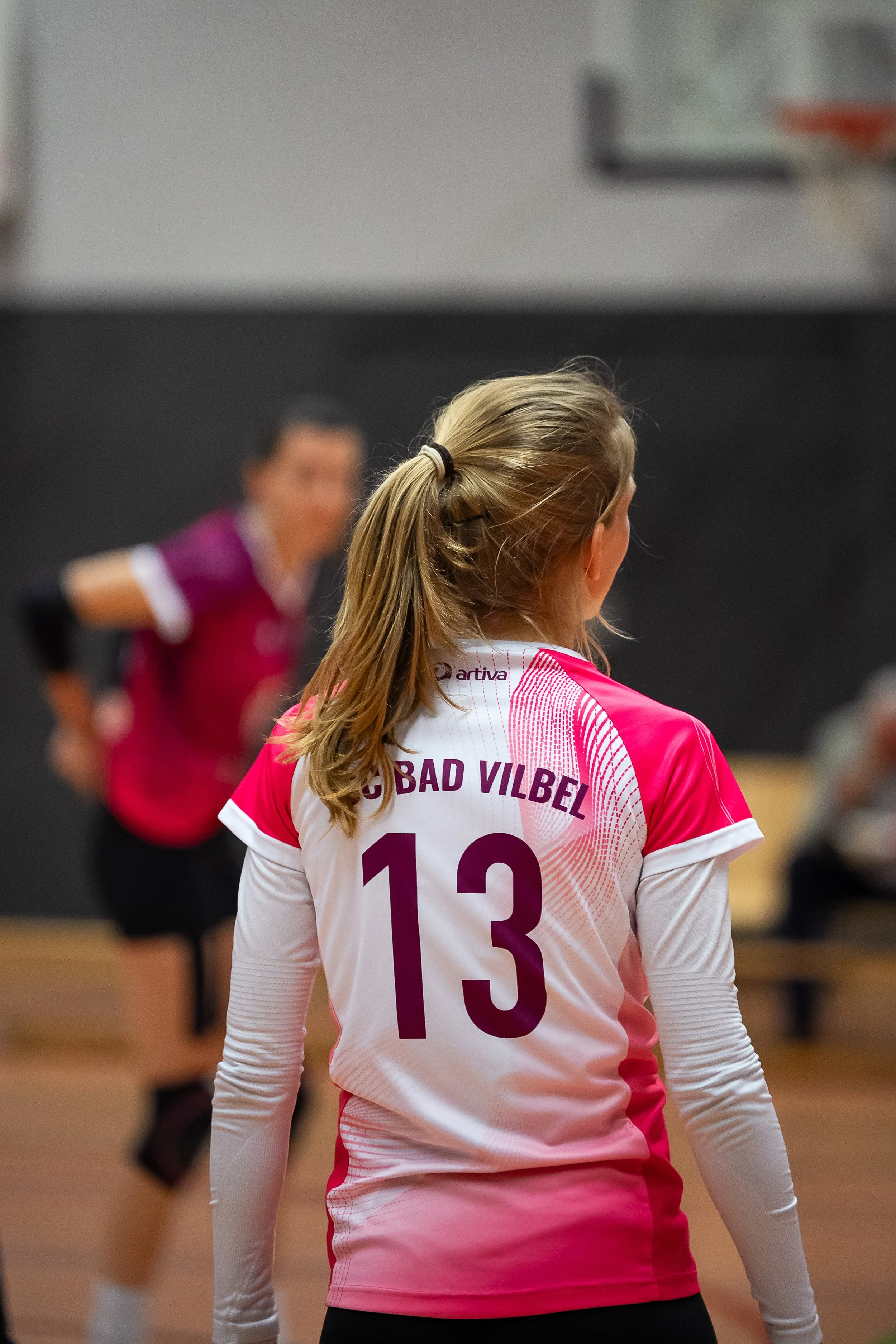 Eine Volleyballspielerin mit der Nummer 13 auf dem Rücken und dem Namen ‚BAD VILBEL‘, die in einem Turnhallen- oder Sportzentrum-Milieu steht, während eine weitere spielende Person im Hintergrund ist.