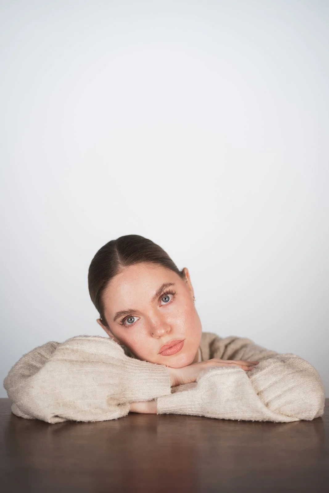 Junge Frau mit braunen Haaren, liegend auf einem Tisch, auf einhelligem weißen Hintergrund, trägt einen beigen Pullover.