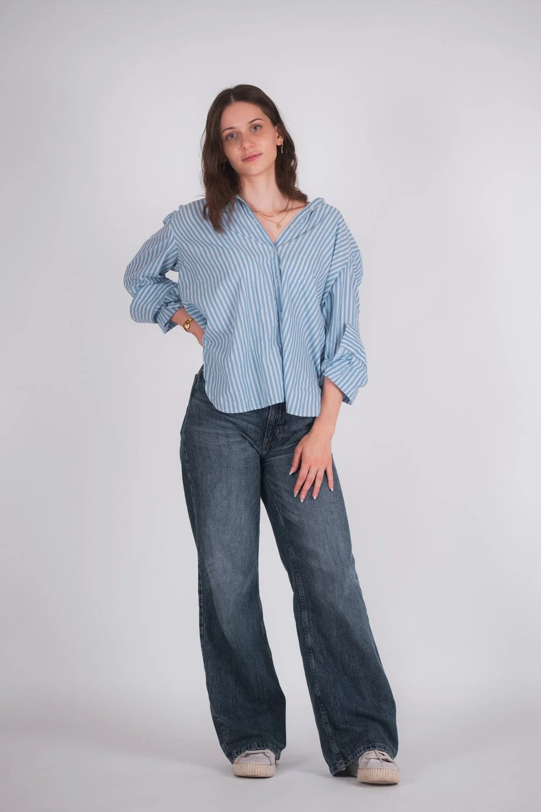 Junge Frau in lockerer blauer gestreifter Bluse, Jeans und Turnschuhe posiert vor hellem Hintergrund.