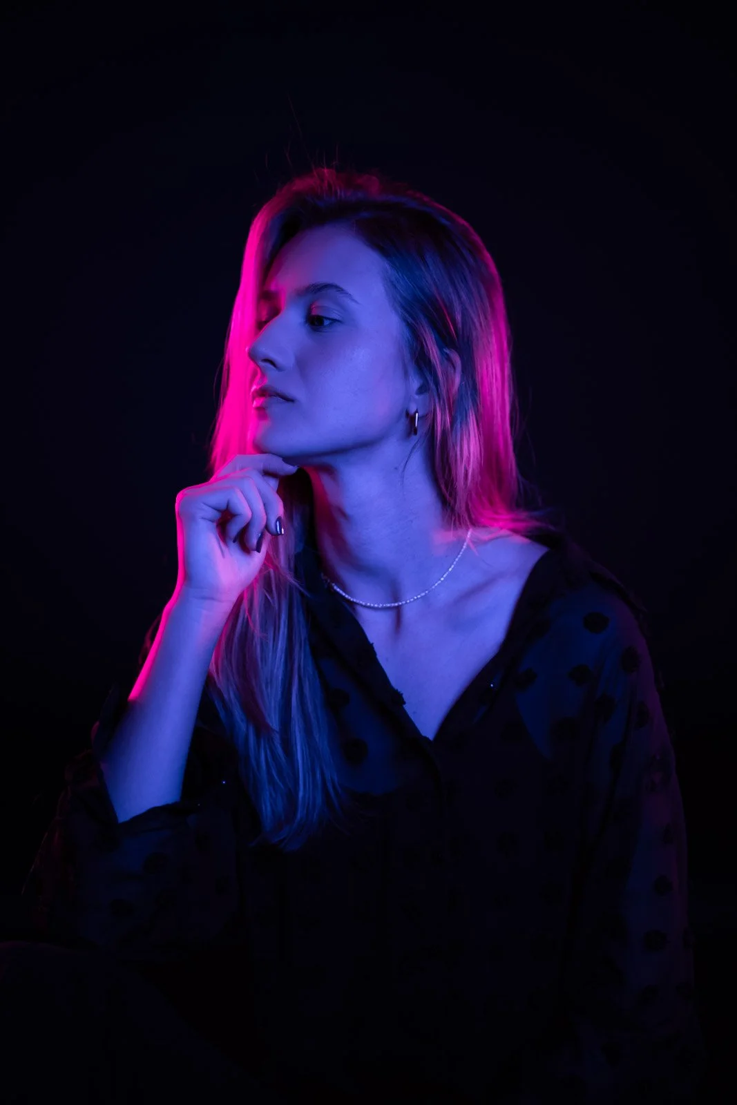 Ein junges Frau sitzt im Dunkeln, beleuchtet von pink und blauen Neonlichtern, trägt eine schwarze Bluse mit Punkten, einen Perlenhalskette und kleine Ohrringe, und hat eine nachdenkliche Haltung.