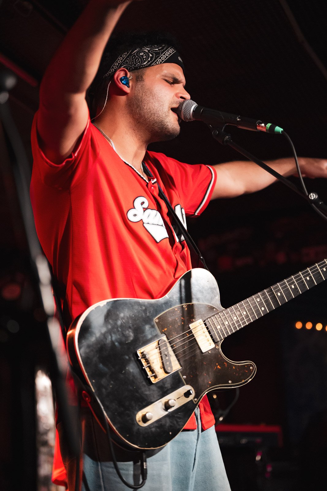 Ein männlicher Musiker trägt ein rotes Trikot, spielt Gitarre, singt in ein Mikrofon, trägt ein schwarzes Stirnband und hat einen blauen Schauspieler-Ohrring, während er auf einer Bühne performt.