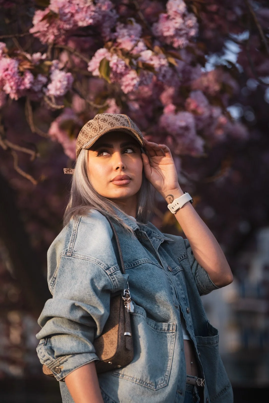 Junge Frau mit grauem Haar in Jeansjacke, Sonnenbrille und Cap, steht vor blühendem Baum, bei Sonnenuntergang.