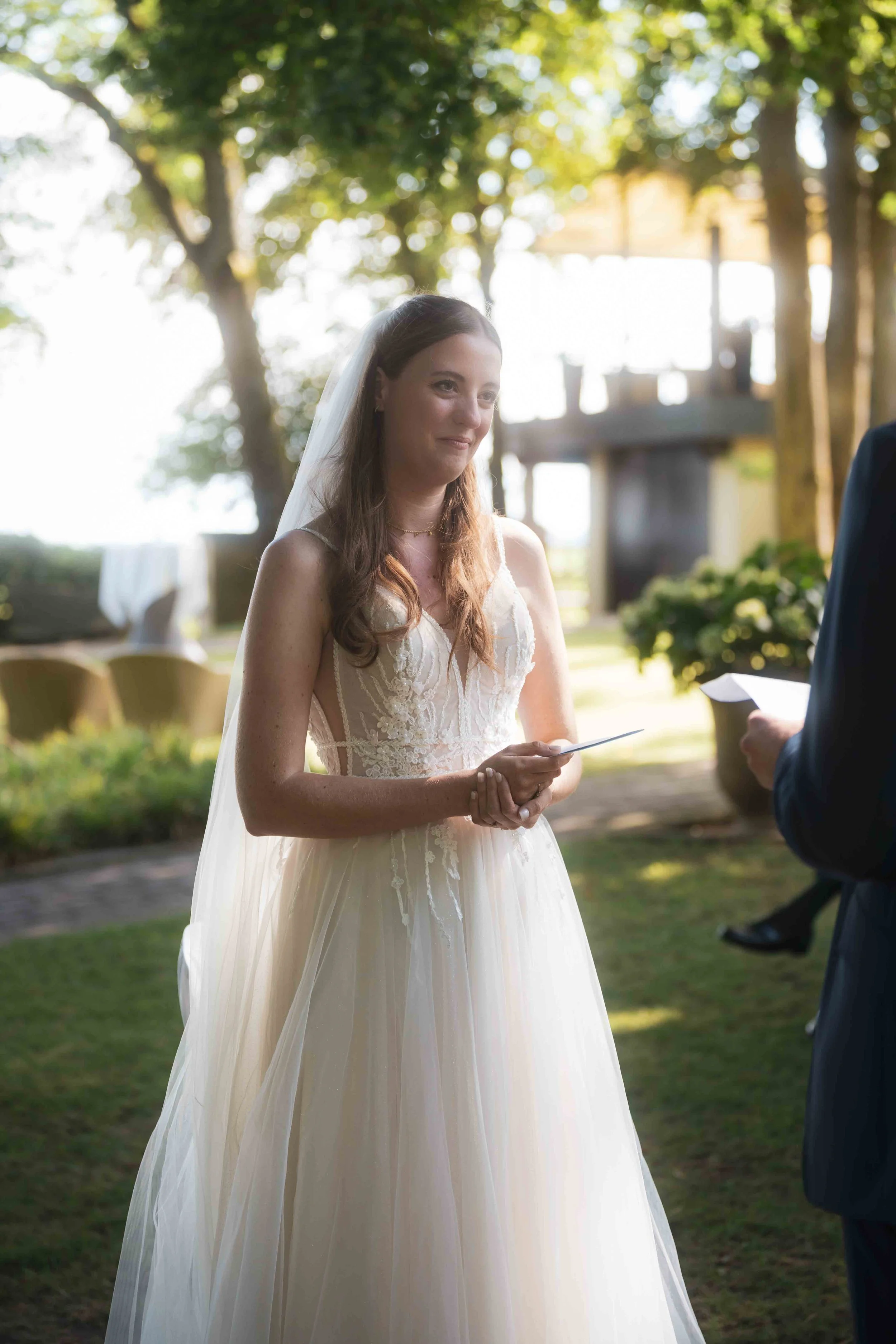Braut im weißen Hochzeitskleid bei einer Trauung im Freien, hält einen Brief oder eine Karte in den Händen, lächelt leicht, im Hintergrund Bäume und ein Gebäude, sonniger Tag.