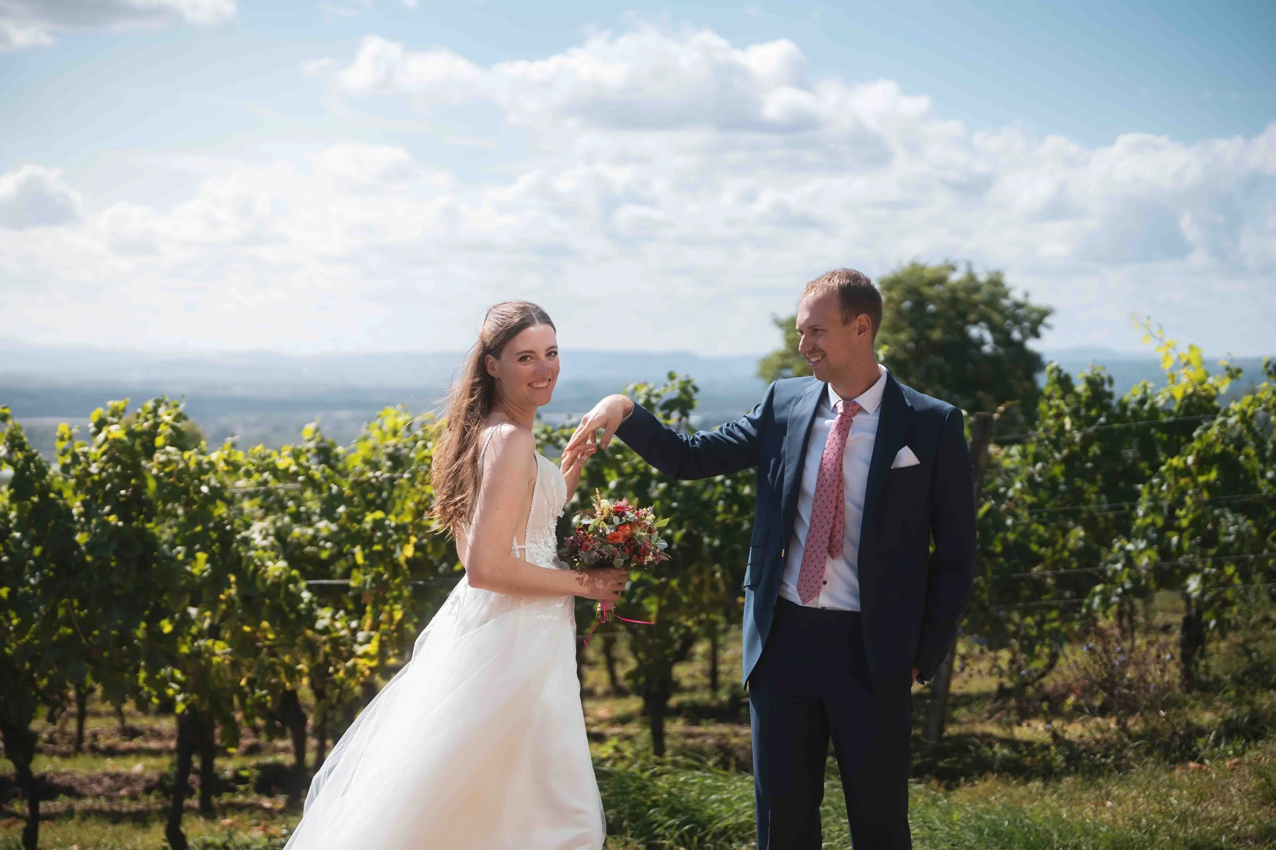 Ein Brautpaar beim Fotoshooting im Weinberg bei sonnigem Himmel, sie lächeln sich an, die Braut trägt ein weißes Hochzeitskleid, der Bräutigam ein dunklen Anzug mit rosa Krawatte, sie hält einen Blumenstrauß.