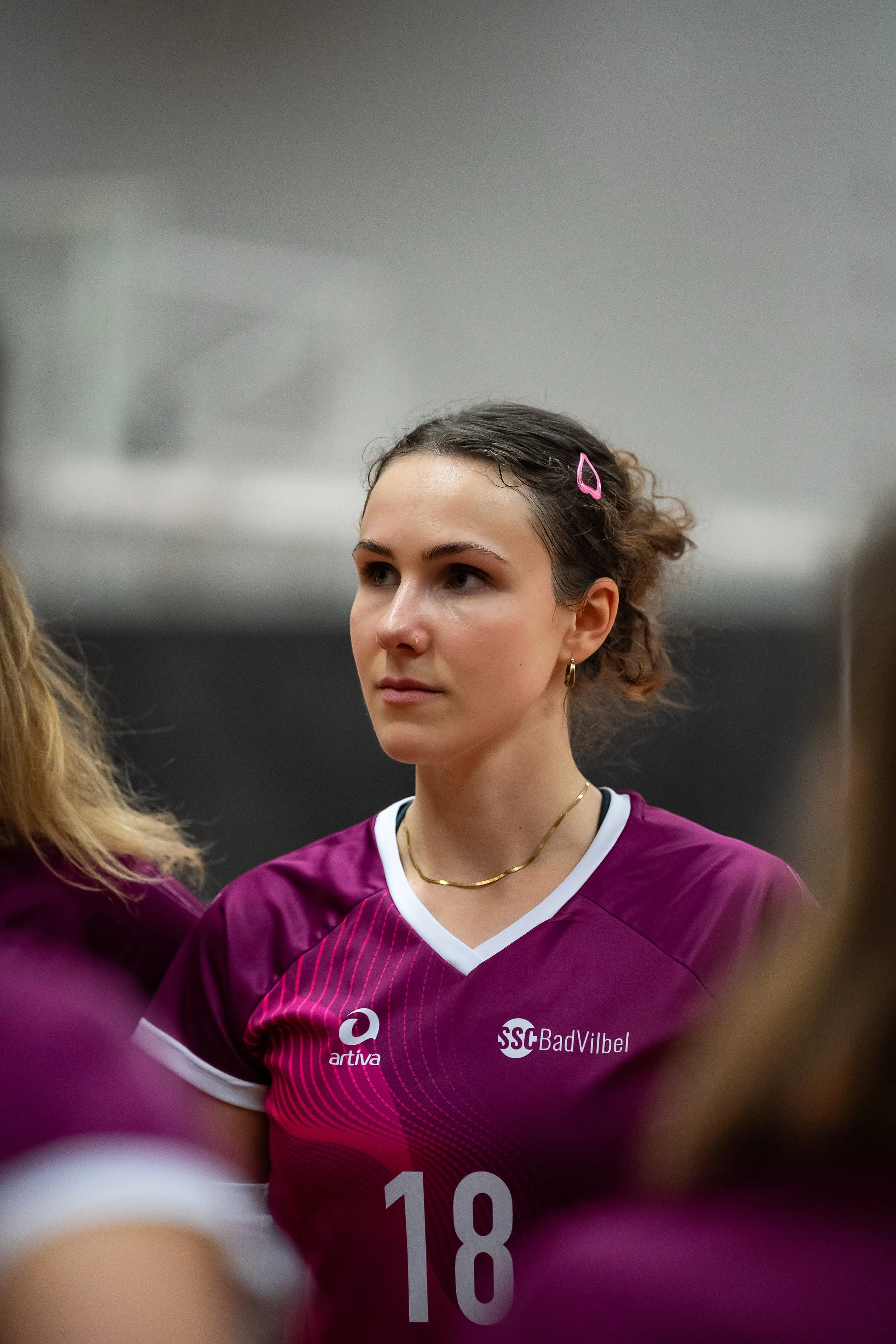 Eine junge Volleyballspielerin in einer burgunderfarbenen Mannschaftsjacke mit der Nummer 18, im Vordergrund, mit dunklen, gewellten Haaren, die mit einem pinken Haarclip zusammengehalten sind.